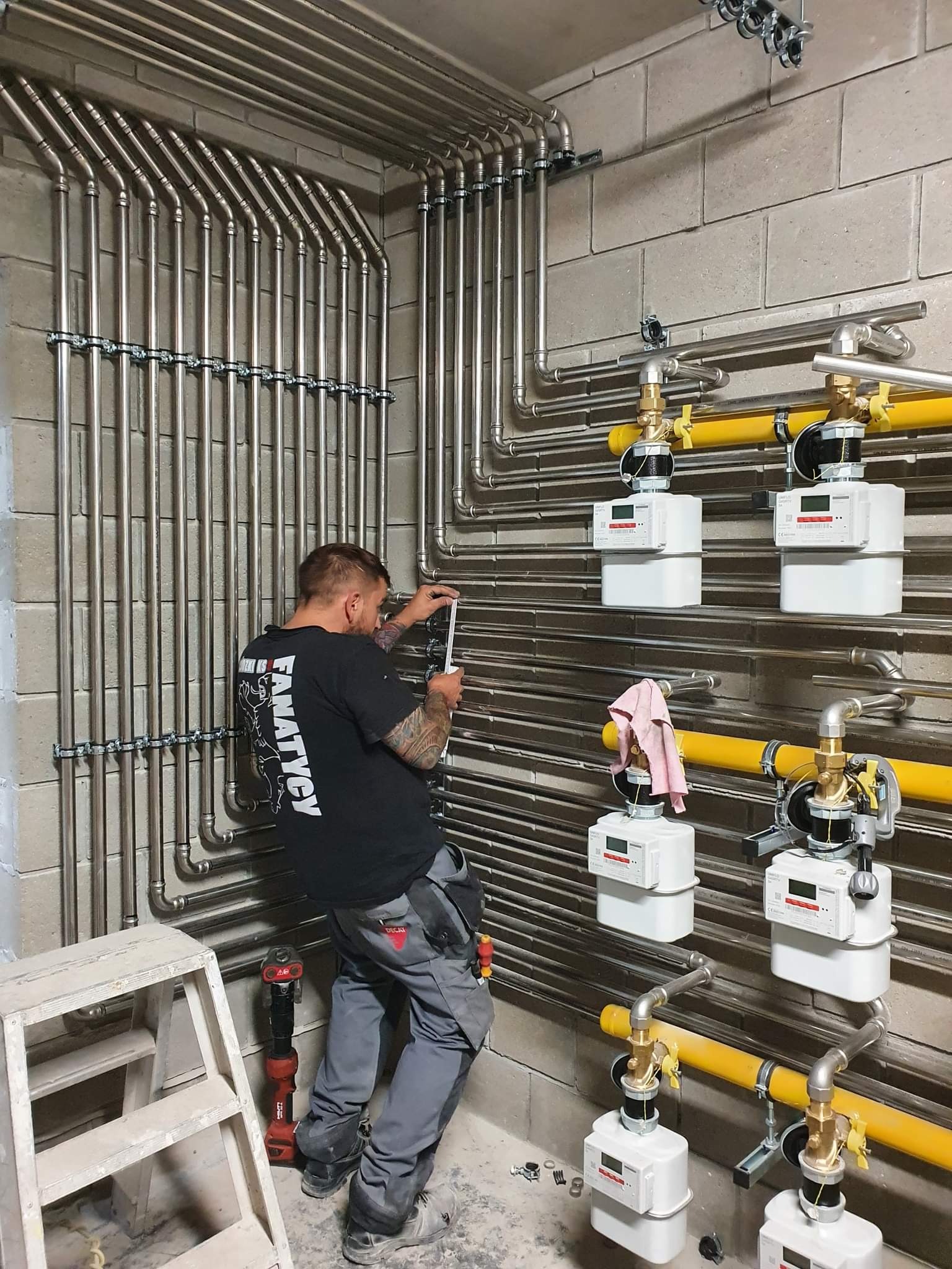 Instalacja gazowa w bloku z szarych cegieł. Hydraulik w czarnej koszulce mierzy rury. Widoczne liczniki gazu, żółte rury i narzędzia pracy.