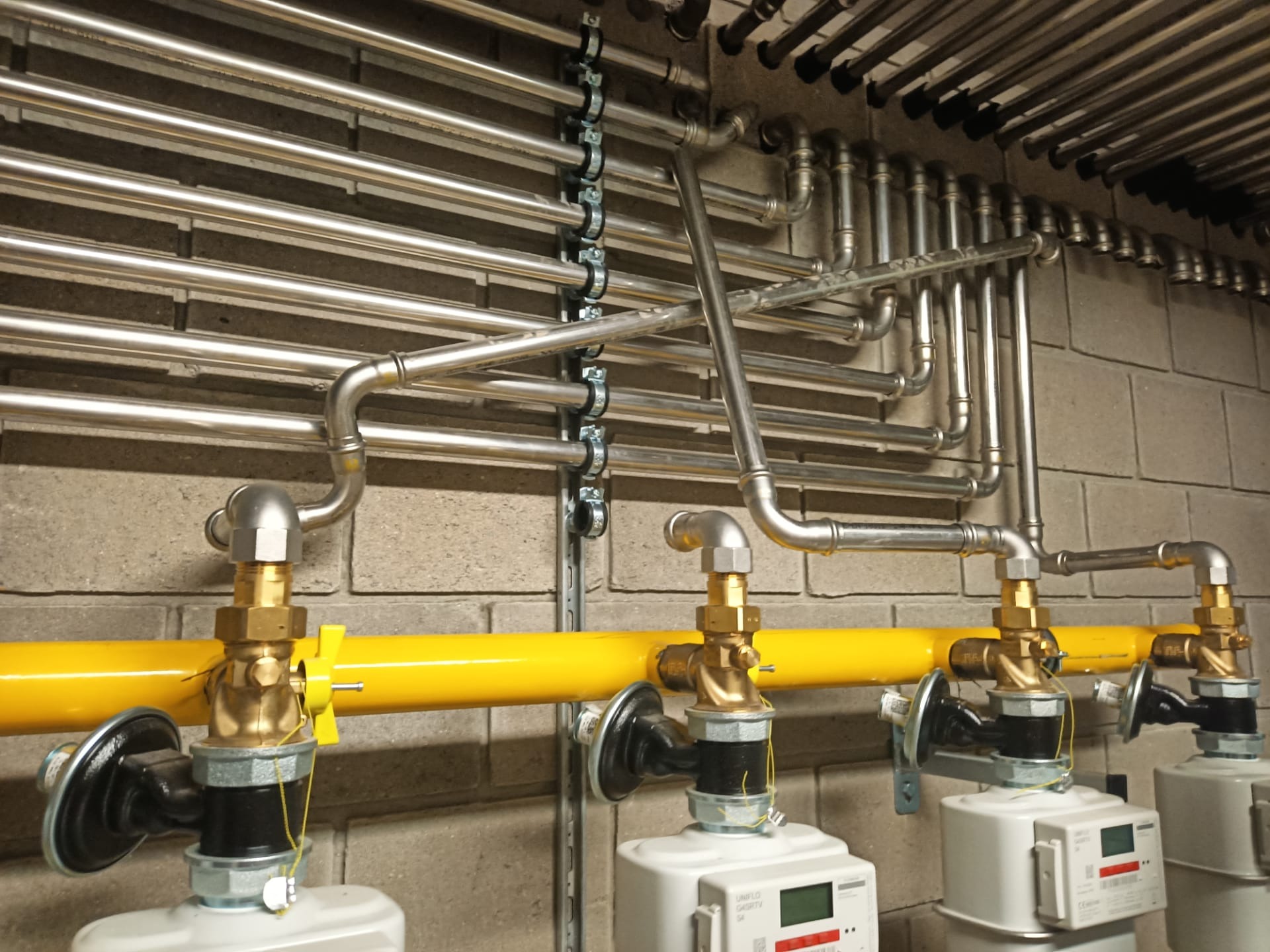 Instalacja gazowa: liczniki, żółta rura i srebrne rury na szarej ścianie z cegieł. Widok z dołu, ukazujący skomplikowaną sieć rur i precyzję wykonania.