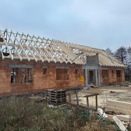 Usługi remontowo- budowlane Mariusz Chojnacki - Surowy stan budynku z czerwonej cegły z drewnianą konstrukcją dachu w trakcie budowy. Widoczne palety i materiały budowlane. Pochmurne niebo w tle.