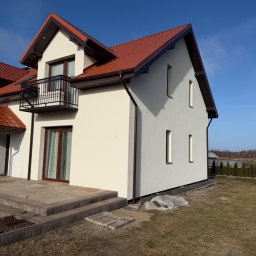 Otynkowany dom jednorodzinny z czerwonym dachem i balkonem, widoczny z boku, na tle błękitnego nieba. Częściowo wykończony taras z kostki brukowej.