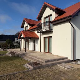 Elewacja domu z balkonem i tarasem wyłożonym kostką. Jasna fasada, czerwony dach i brązowe rynny. Widok na zadbany ogród i błękitne niebo.