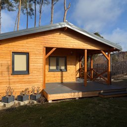 Drewniany domek letniskowy z zadaszonym tarasem i szarymi oknami, otoczony zielenią i drzewami iglastymi, w słoneczny dzień. Widoczne donice z roślinami.