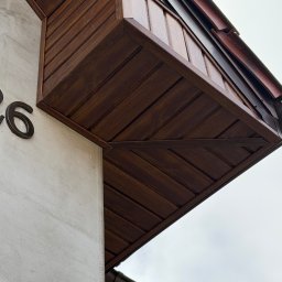 Detal elewacji budynku z drewnianą obudową okapu w ciepłym brązie, numer domu '26' na jasnej, teksturowanej ścianie. Ujęcie z dołu, widoczne fragmenty rynny.