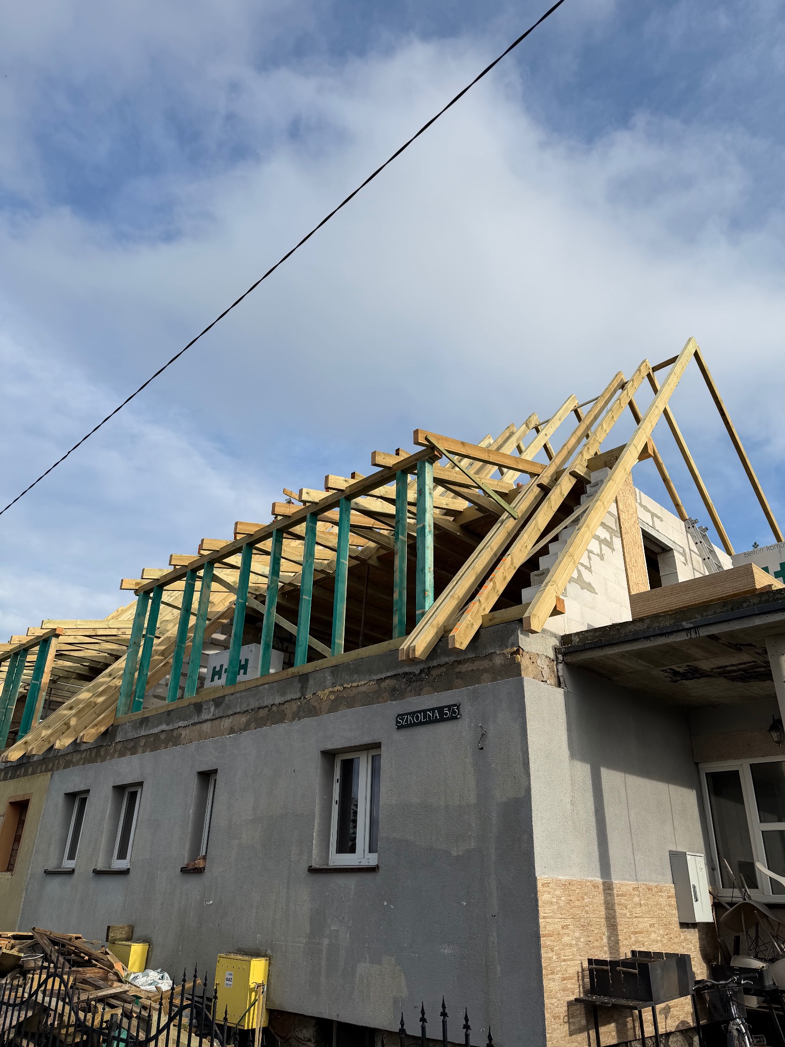 Budowa dachu domu jednorodzinnego w trakcie realizacji. Widoczne drewniane elementy konstrukcyjne i fragmenty ścian. Błękitne niebo w tle.