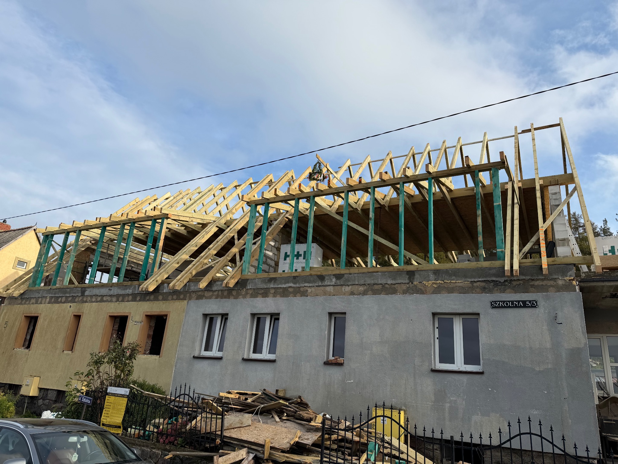 Budowa dachu na dwukondygnacyjnym budynku z widoczną konstrukcją drewnianą i stertą materiałów budowlanych przed budynkiem. Widoczne niebo z chmurami.