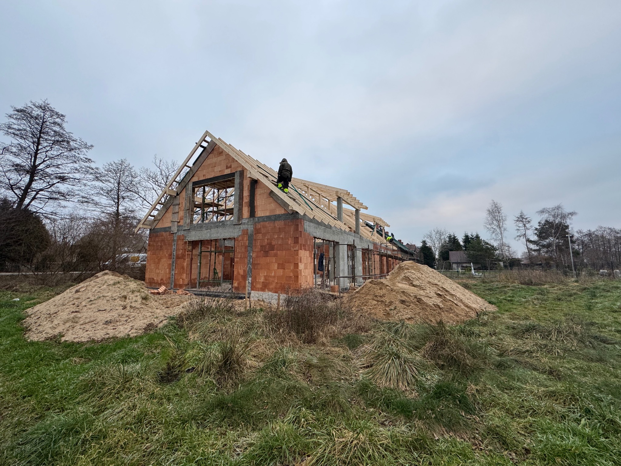 Budowa domu jednorodzinnego w stanie surowym otwartym. Widoczne mury z czerwonej cegły, drewniana konstrukcja dachu i pracownicy na górze. Dwie pryzmy piasku na pierwszym planie.