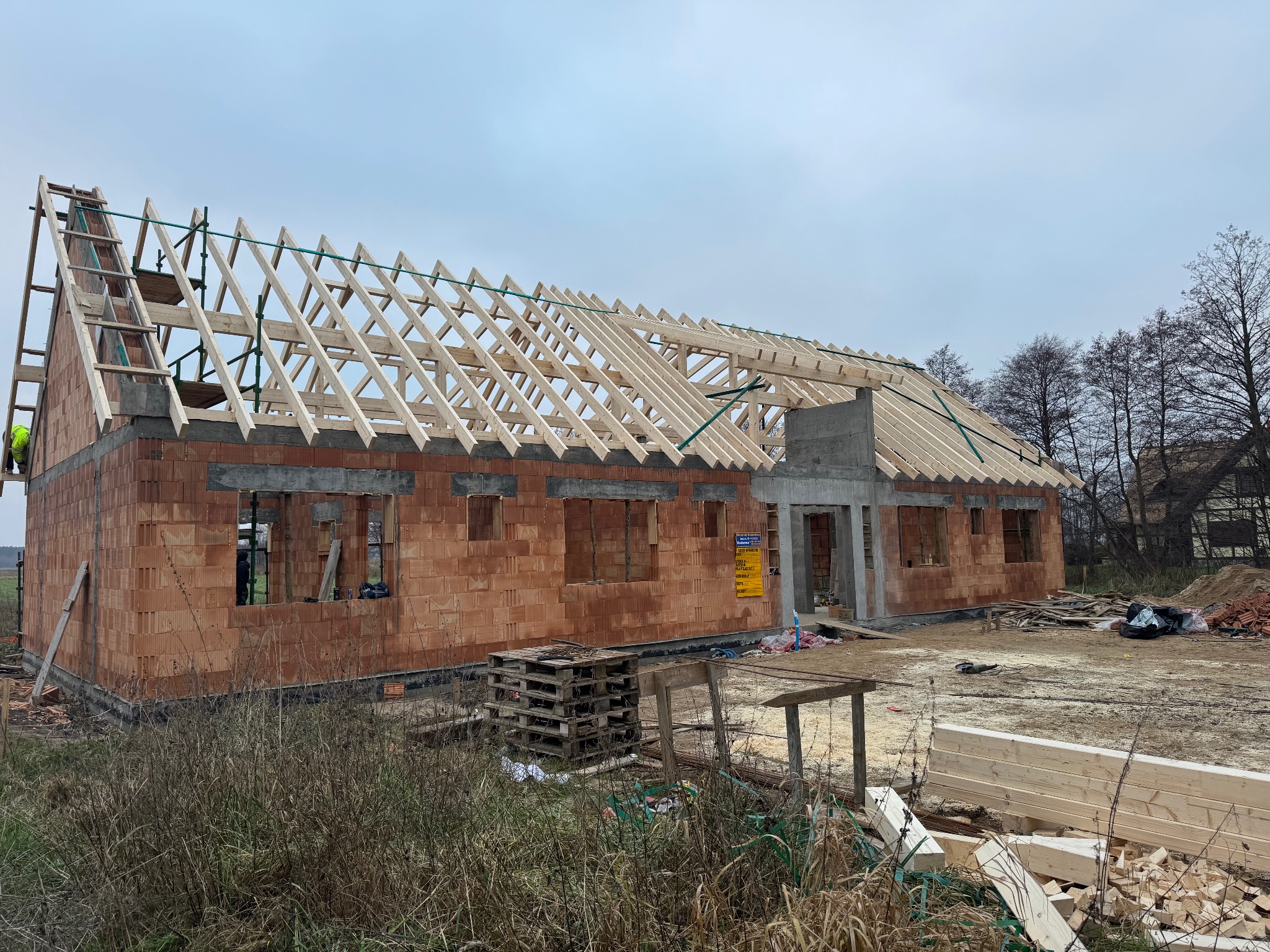 Surowy stan budynku z czerwonej cegły z drewnianą konstrukcją dachu w trakcie budowy. Widoczne palety i materiały budowlane. Pochmurne niebo w tle.