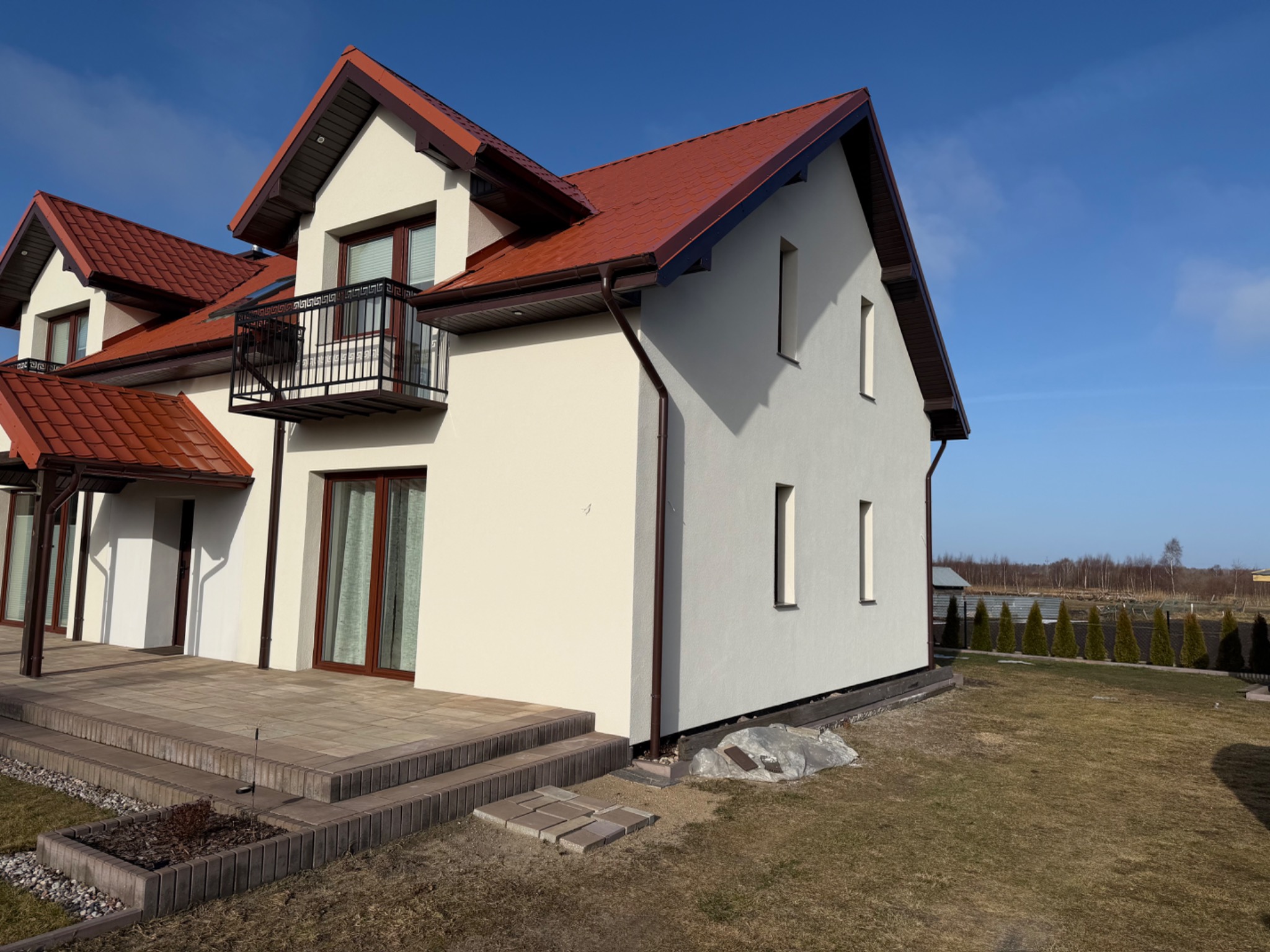 Otynkowany dom jednorodzinny z czerwonym dachem i balkonem, widoczny z boku, na tle błękitnego nieba. Częściowo wykończony taras z kostki brukowej.