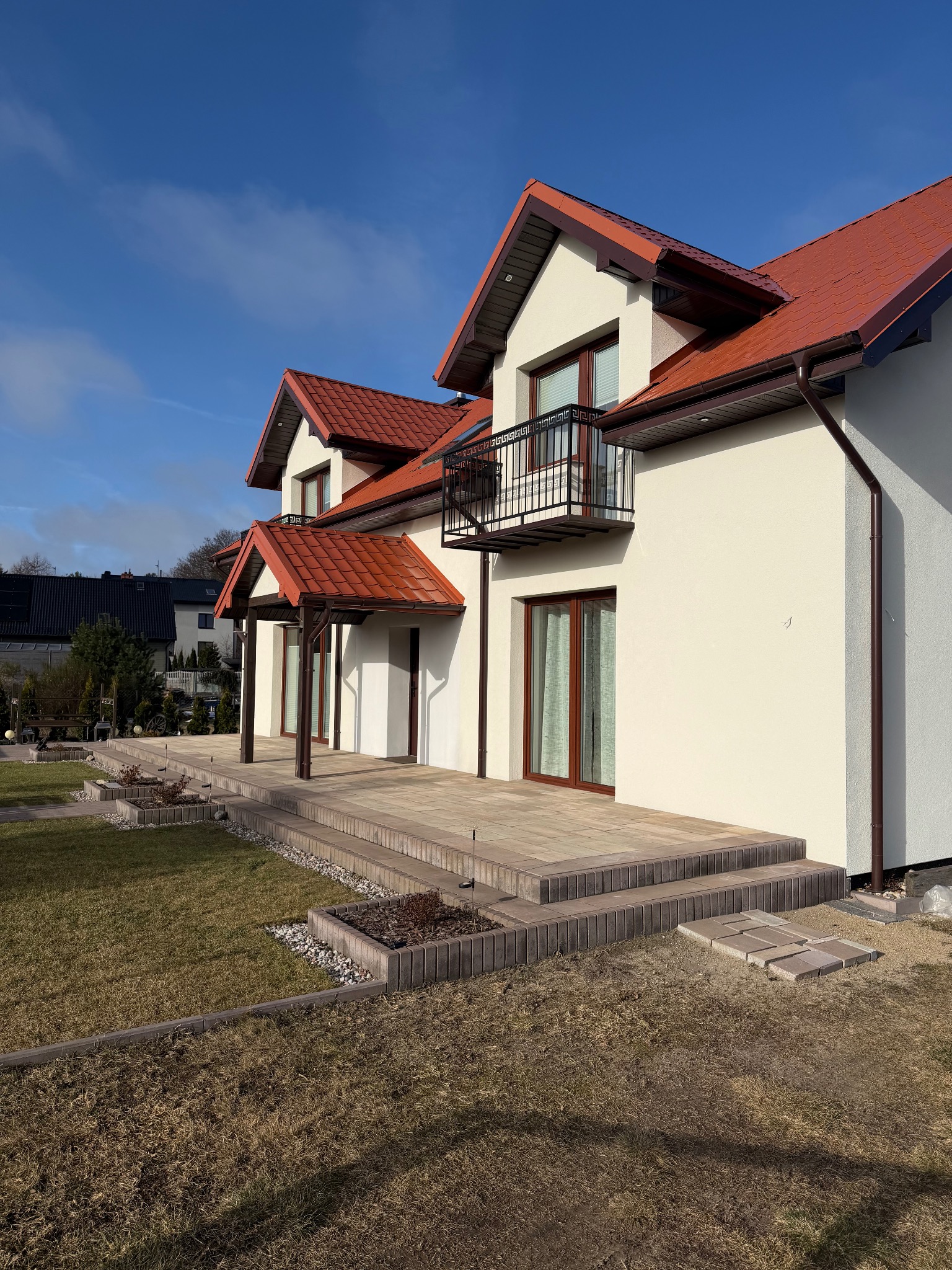Elewacja domu z balkonem i tarasem wyłożonym kostką. Jasna fasada, czerwony dach i brązowe rynny. Widok na zadbany ogród i błękitne niebo.