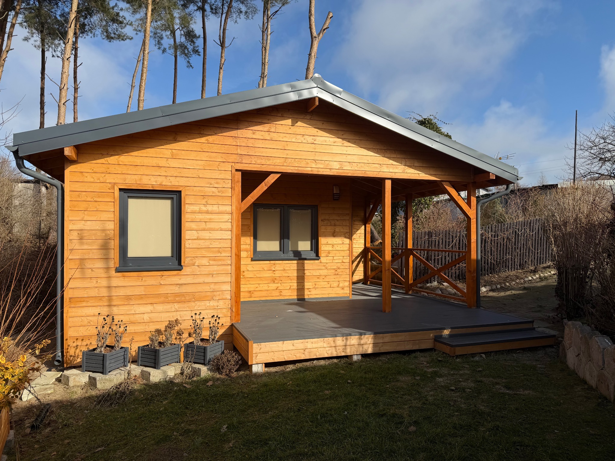 Drewniany domek letniskowy z zadaszonym tarasem i szarymi oknami, otoczony zielenią i drzewami iglastymi, w słoneczny dzień. Widoczne donice z roślinami.