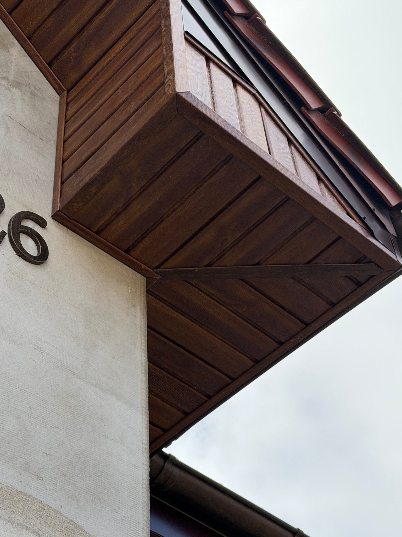 Detal elewacji budynku z drewnianą obudową okapu w ciepłym brązie, numer domu '26' na jasnej, teksturowanej ścianie. Ujęcie z dołu, widoczne fragmenty rynny.