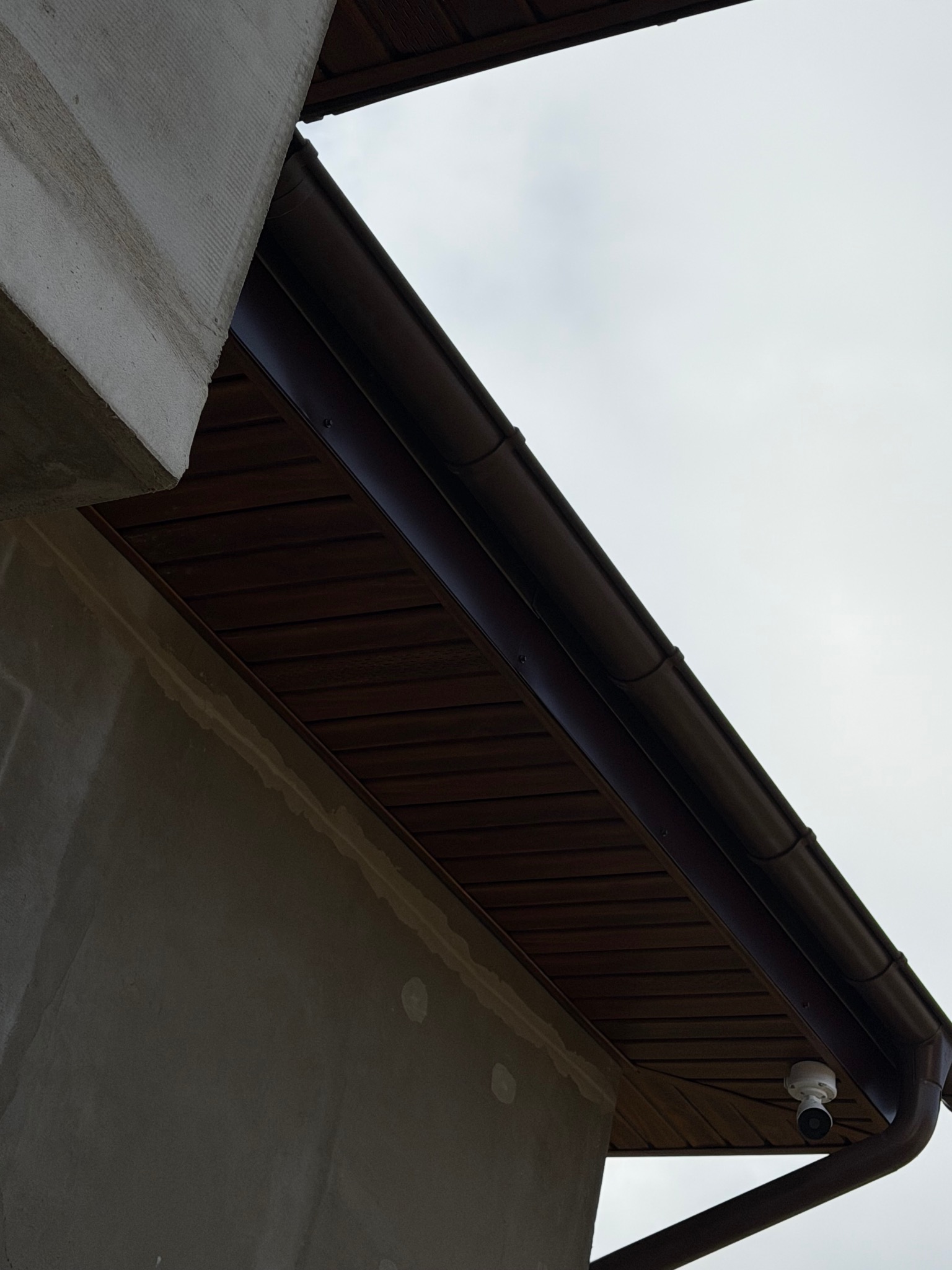 Fragment elewacji budynku z brązową podbitką i rynną, widoczna kamera monitoringu. Ściana w stanie surowym, przygotowana do dalszych prac wykończeniowych.
