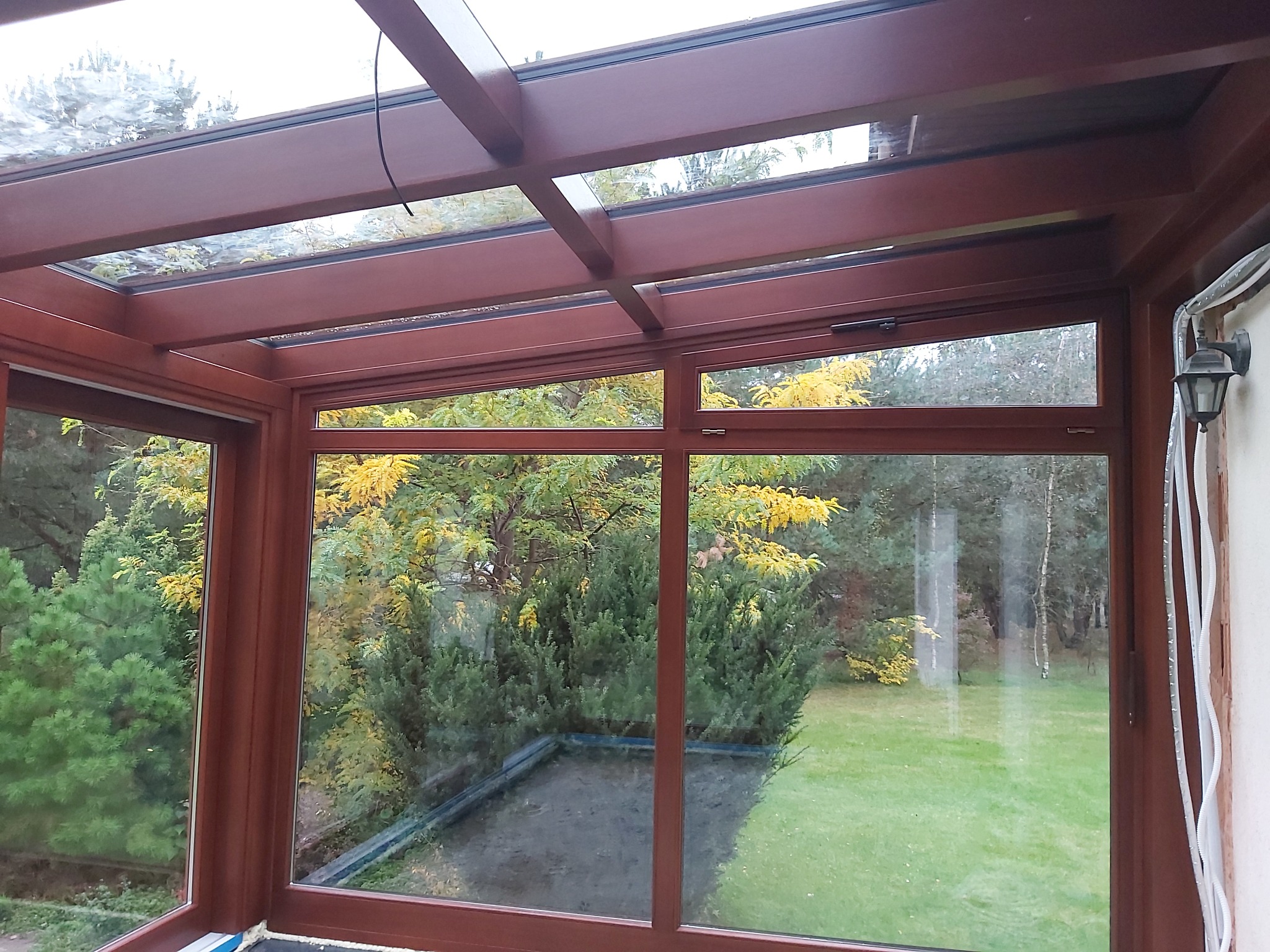 Zadaszony taras z brązowymi ramami okiennymi i szklanym dachem, widok na zieleń ogrodu. Charakterystyczne oświetlenie zewnętrzne i widoczne przewody elektryczne.