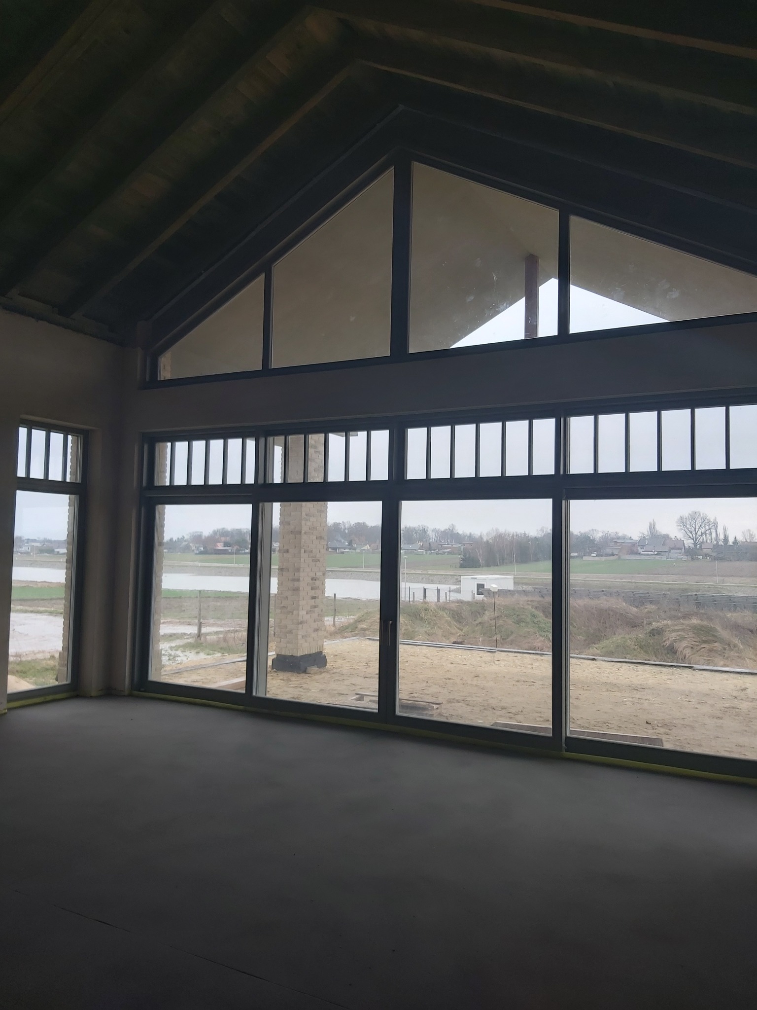 Duże, panoramiczne okna z czarnymi ramami w surowym wnętrzu domu w trakcie budowy. Widok na krajobraz za oknem. Charakterystyczny kształt okien w szczycie dachu.