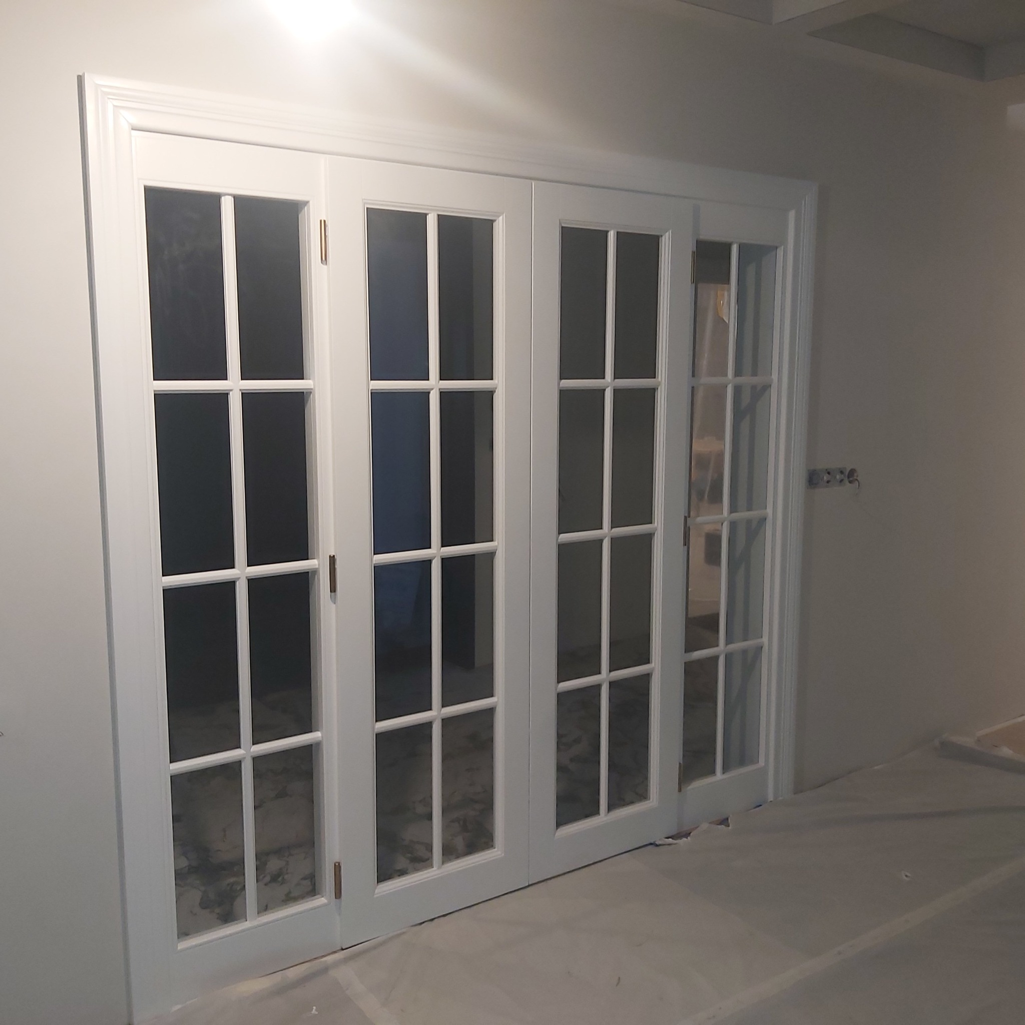 Białe, przeszklone drzwi dwuskrzydłowe z podziałami, widoczne na tle jasnej ściany. Podłoga zabezpieczona folią malarską. Minimalistyczny design.