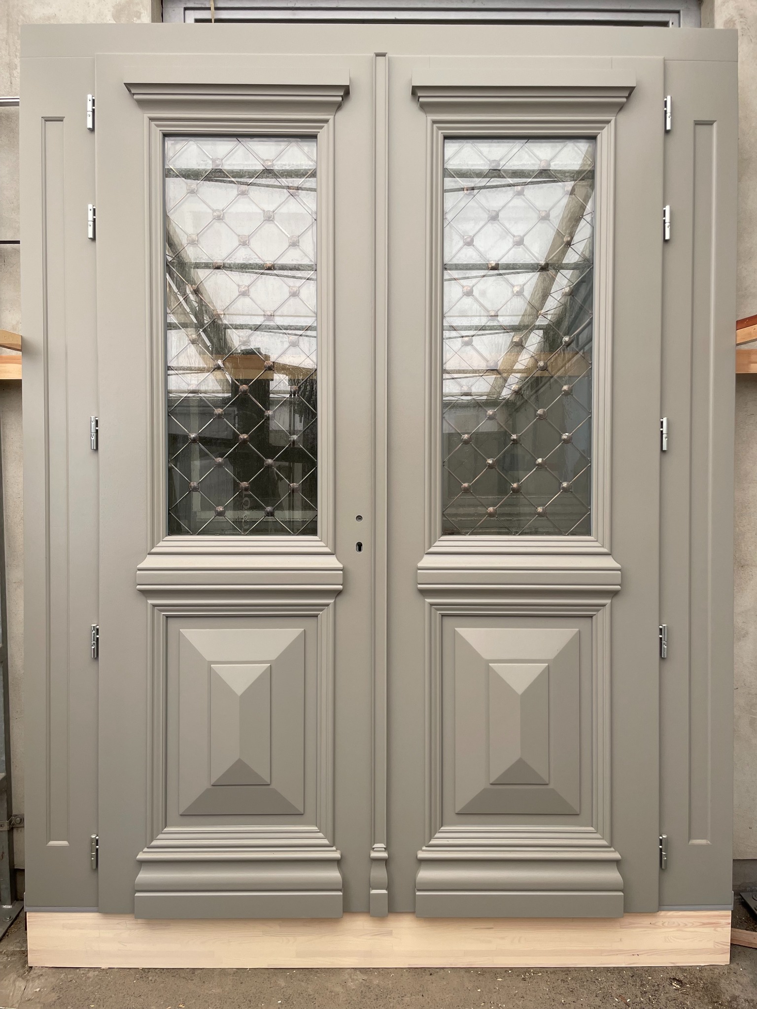 Szare, dwuskrzydłowe drzwi z szybami w kratkę, ozdobnymi panelami i srebrnymi zawiasami, ustawione na drewnianych legarach. Widoczne odbicie konstrukcji dachu.