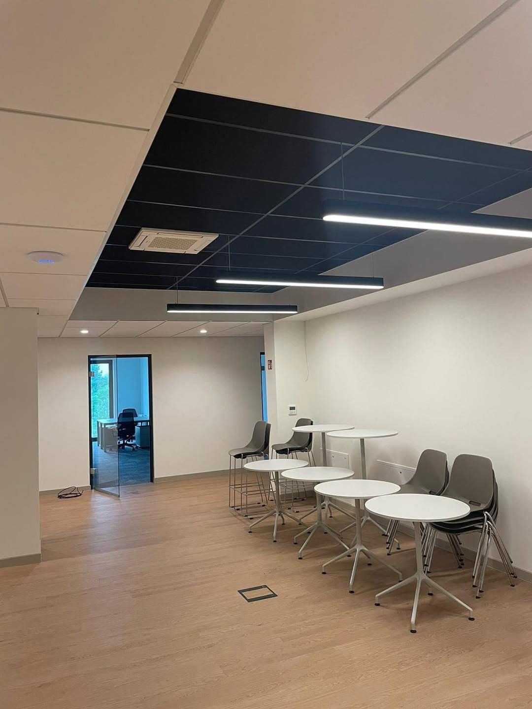 Nowoczesne biuro po remoncie: podwieszany sufit z płyt karton-gips, oświetlenie liniowe, jasne ściany i podłoga. Widoczne stoliki i krzesła.