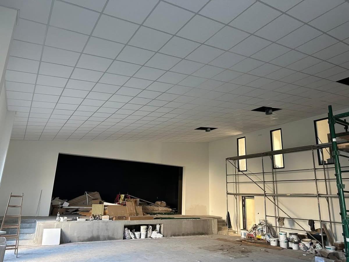 Sufit podwieszany z płyt, kratownica z widocznymi otworami wentylacyjnymi, ściany pomalowane na biało, rusztowanie i scena w tle. Prace wykończeniowe w dużej sali.