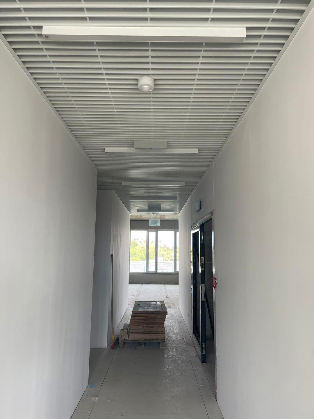 Korytarz w trakcie prac wykończeniowych: ściany przygotowane pod malowanie, sufit podwieszany z kratką, widoczne płyty na podłodze i otwarte drzwi.