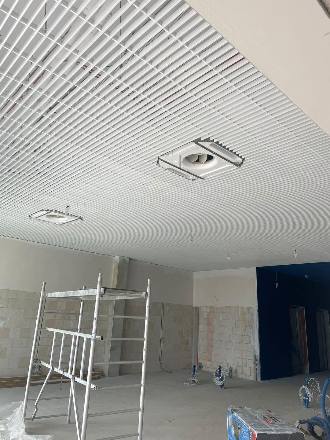 Biały sufit rastrowy z kratką i wbudowanymi elementami wentylacji, widok z dołu, rusztowanie w pomieszczeniu w trakcie prac wykończeniowych.