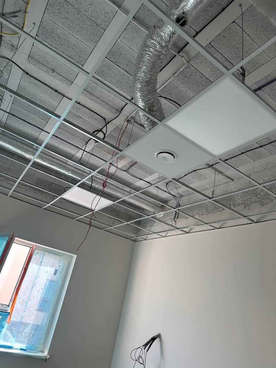 Sufit podwieszany z widoczną konstrukcją metalową, instalacją wentylacyjną i oświetleniem panelowym w nowym pomieszczeniu z oknem. Widok z dołu.