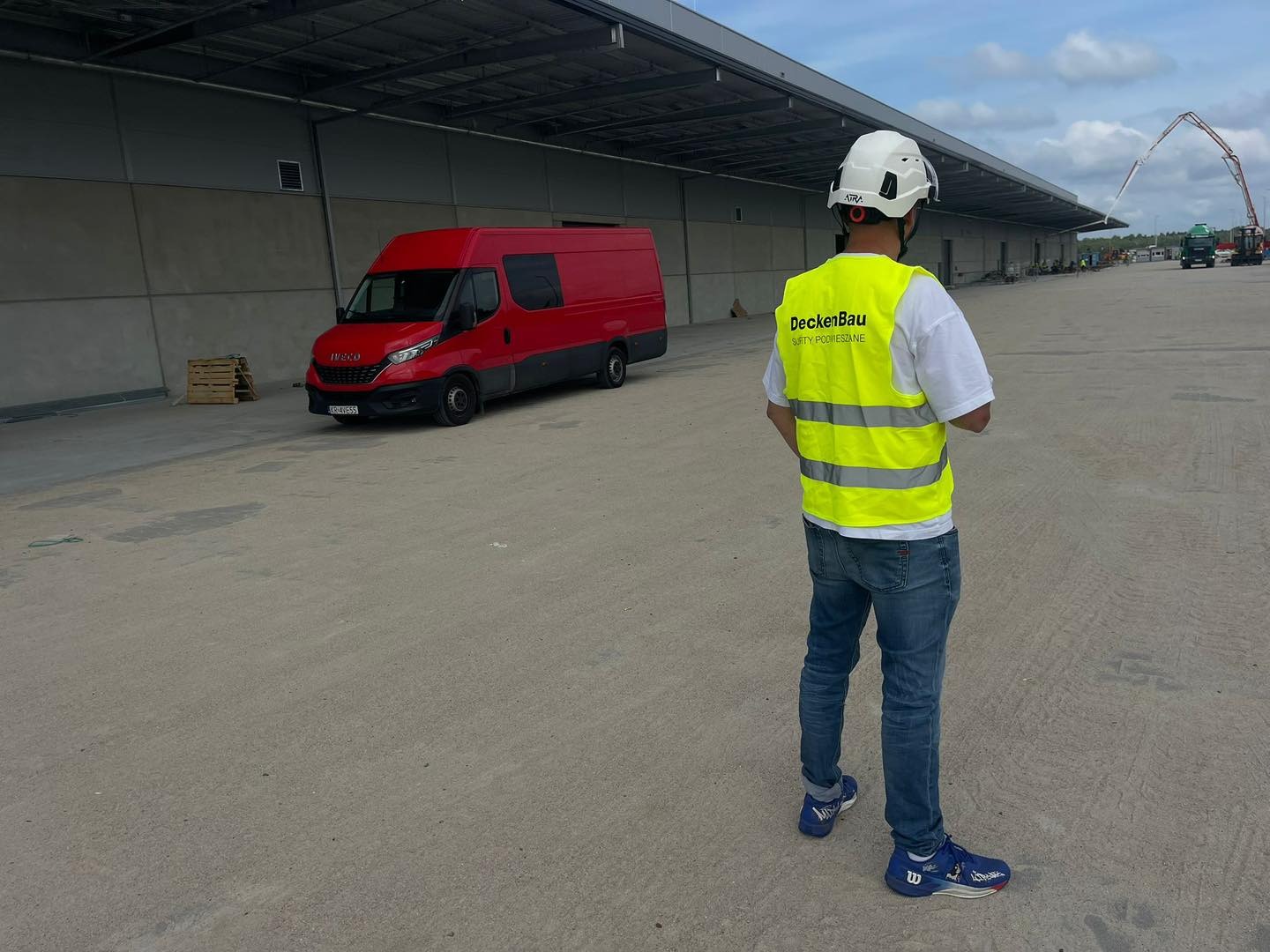 Pracownik w kamizelce odblaskowej i kasku na tle hali, czerwonego busa i pompy do betonu. Widok z tyłu, skupienie na detalu kamizelki z nazwą firmy.