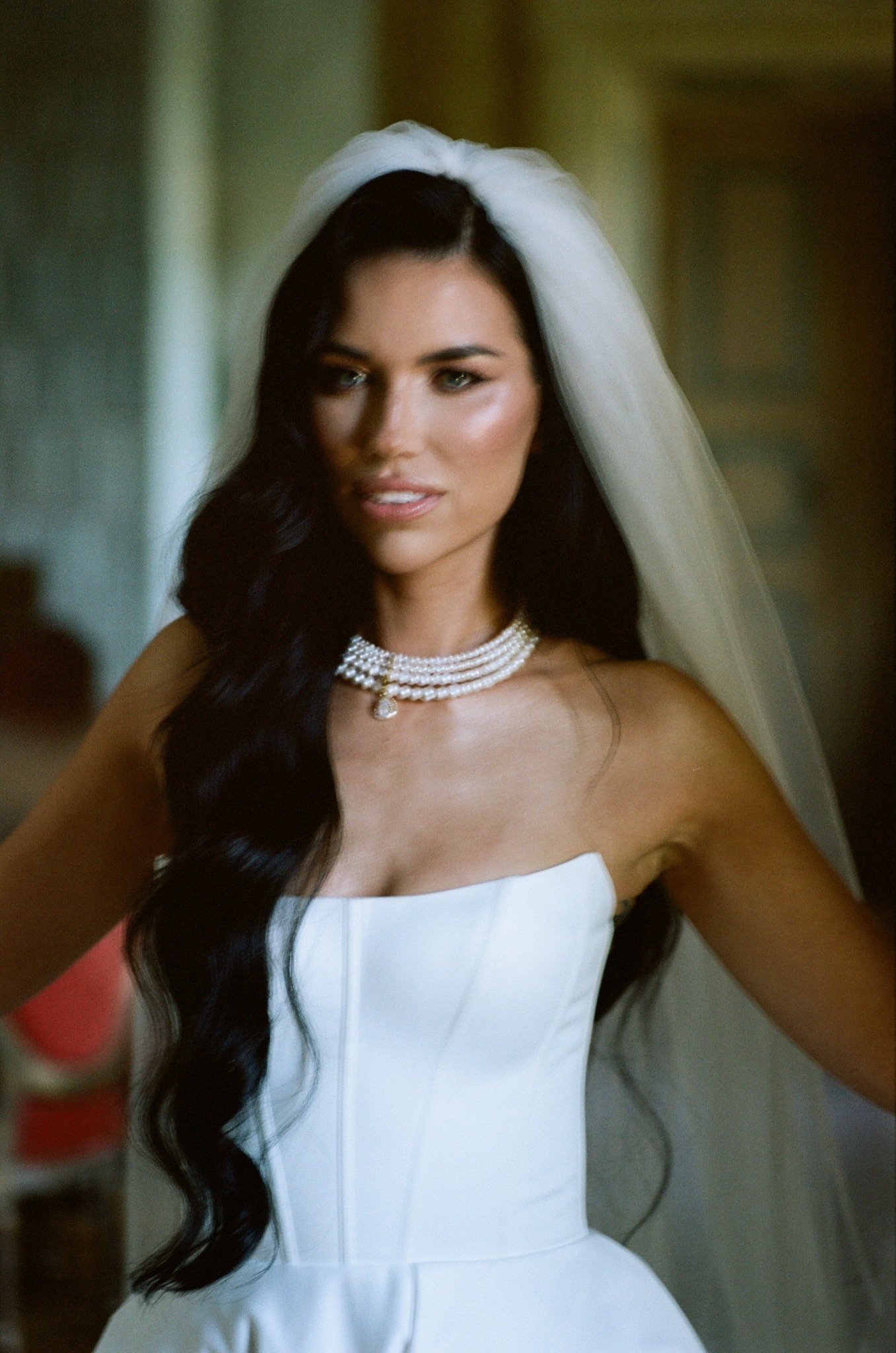 Portret kobiety w białej sukni ślubnej z welonem i perłowym naszyjnikiem, uśmiechająca się, z ciemnymi, falowanymi włosami. Delikatne oświetlenie, subtelny makijaż.