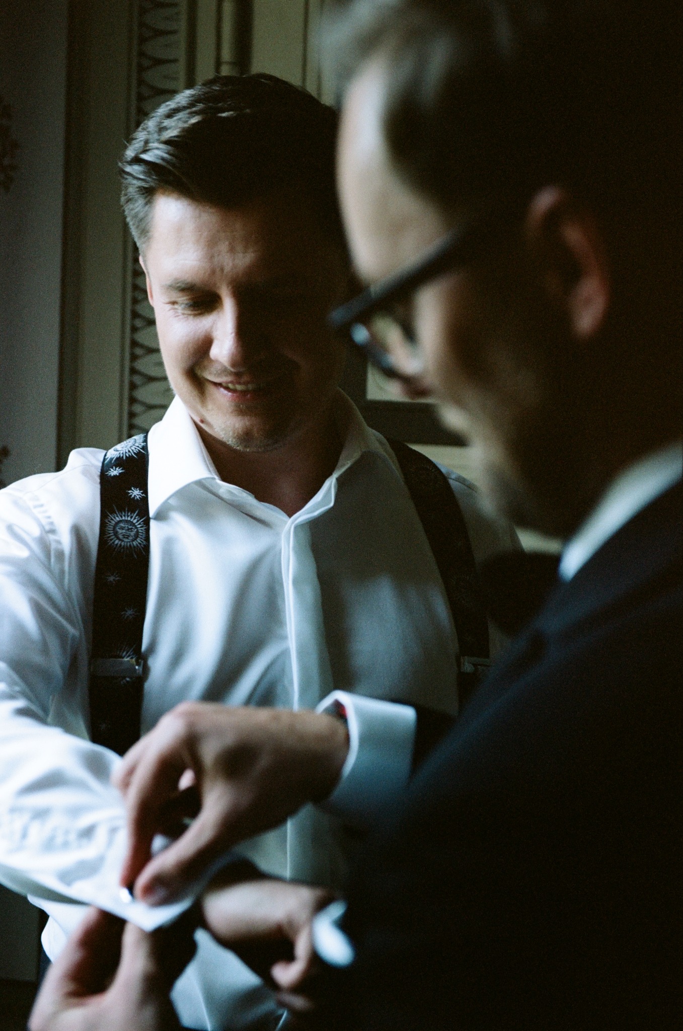 Uśmiechnięty mężczyzna w białej koszuli i szelkach zapina mankiet, drugi mężczyzna w garniturze obserwuje. Przygotowania do ślubu w klimatycznym wnętrzu.