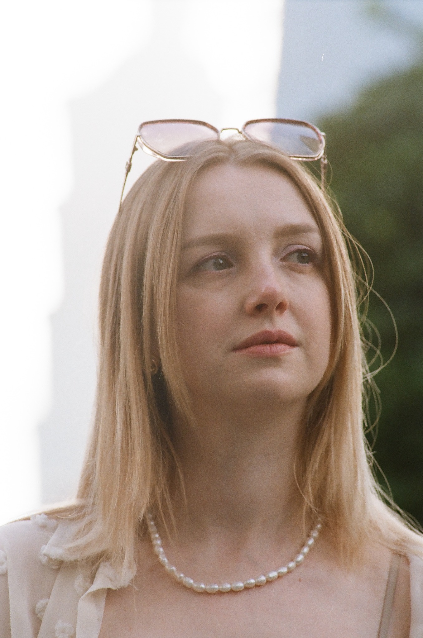 Portret kobiety z blond włosami, w perłowym naszyjniku i okularami na głowie, spoglądającej w górę, w naturalnym świetle.