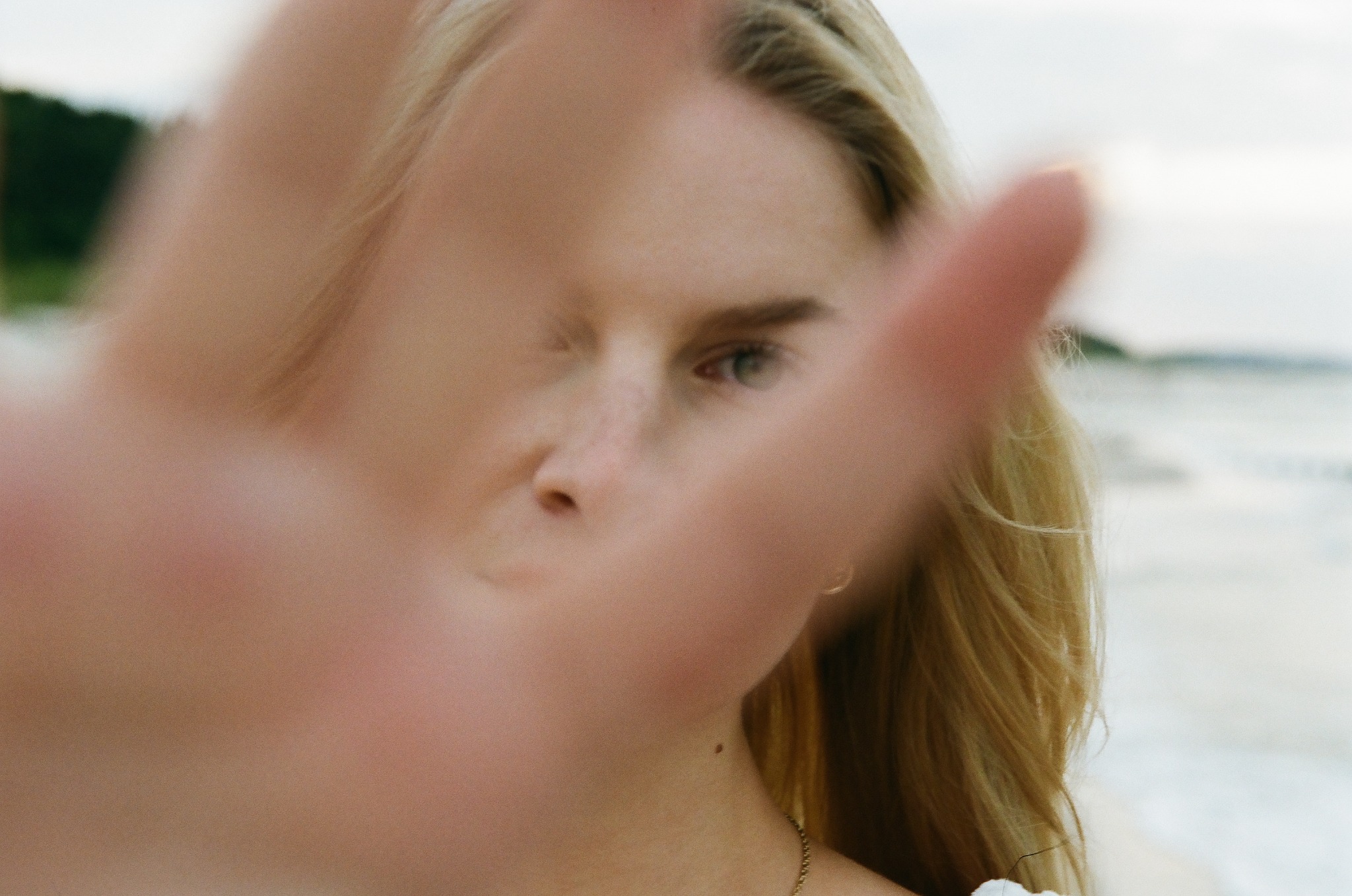 Portret kobiety z blond włosami, częściowo zasłoniętej dłonią, na tle plaży. Ujęcie z bliska, skupione na twarzy i ekspresji.