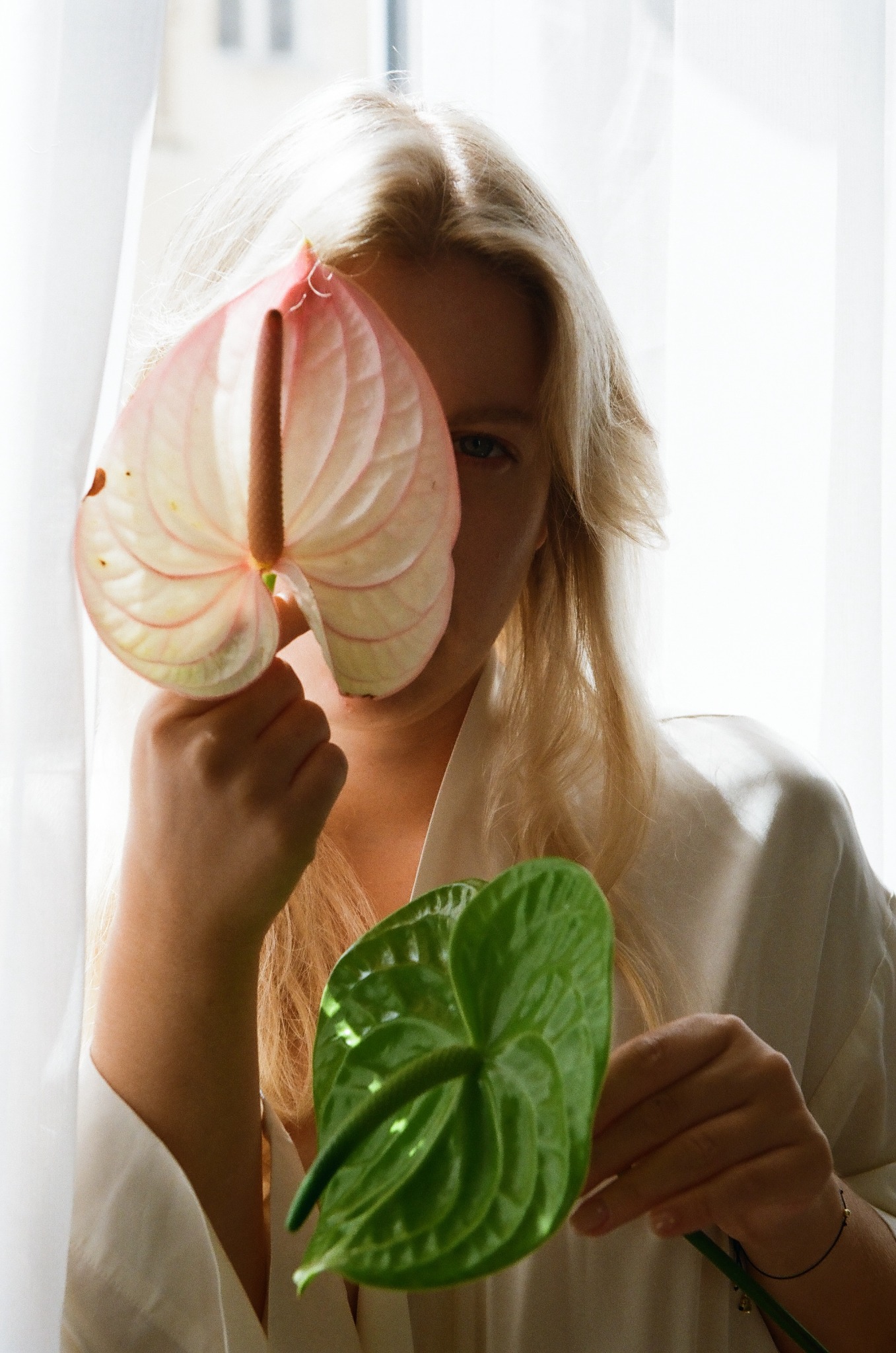 Blondynka w bieli, z anturium w dłoniach na tle okna. Jeden kwiat różowy, drugi zielony, zasłaniają częściowo twarz. Delikatne światło, subtelny makijaż.