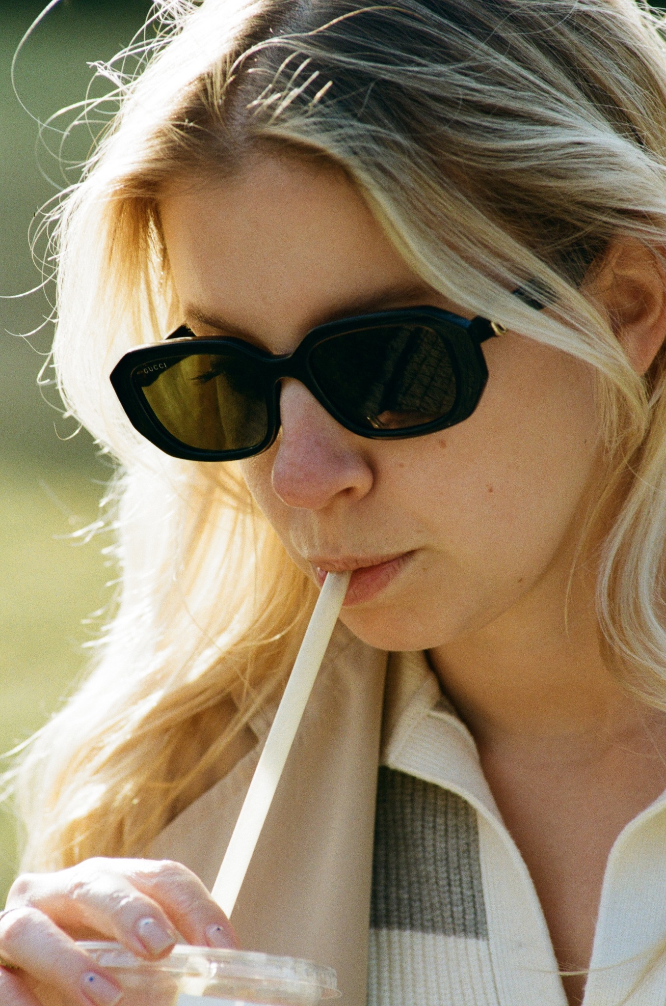 Portret kobiety w okularach Gucci, pijącej napój przez słomkę. Jasne włosy, delikatny makijaż, naturalne światło. Ujęcie z bliska, skupione na twarzy i detalach.