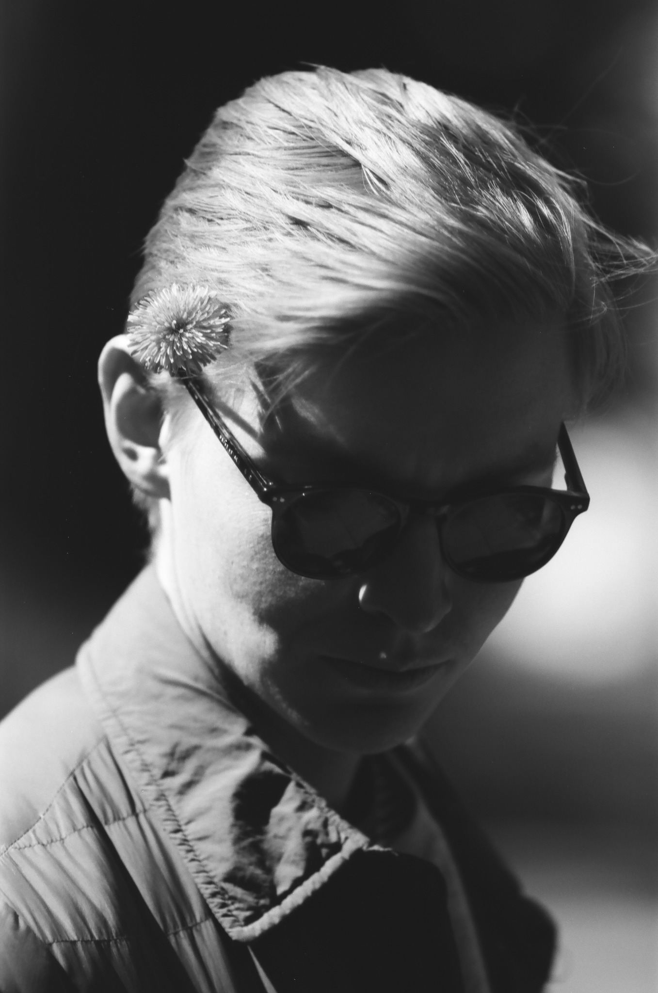 Czarno-biały portret kobiety w okularach, z dmuchawcem za uchem, spoglądającej w dół, w miękkim świetle.