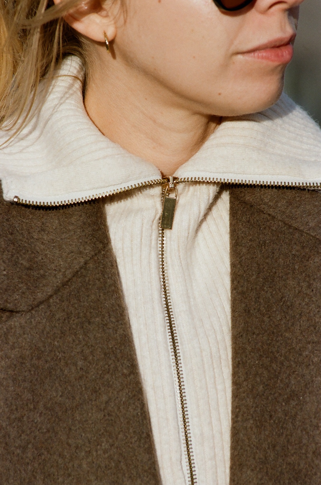 Fragment portretu kobiety w okularach przeciwsłonecznych, ubrana w sweter z zamkiem i brązową marynarkę. Widoczny złoty kolczyk w uchu.