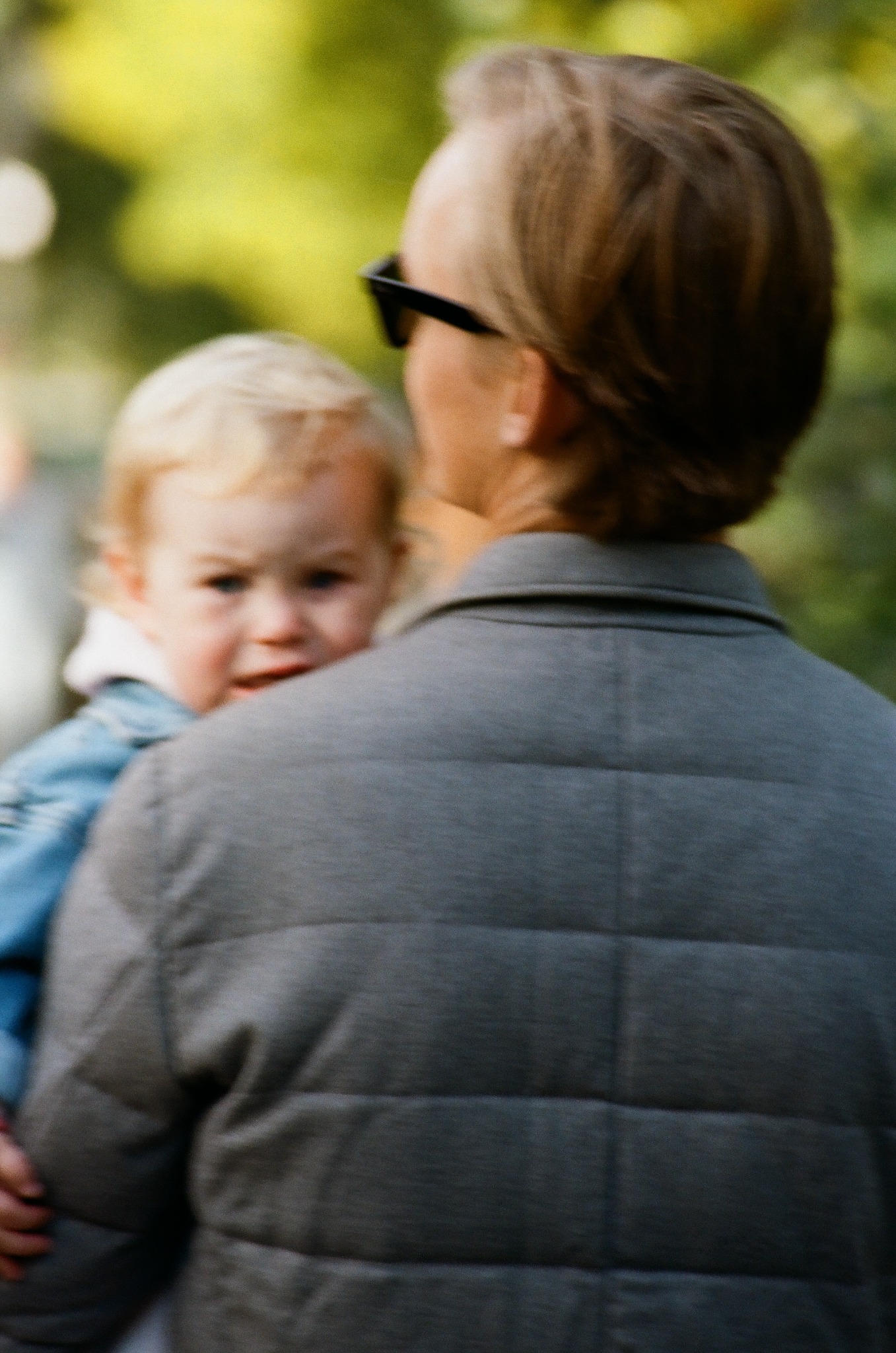 Blondwłosa dziewczynka spogląda przez ramię osoby dorosłej w ciemnych okularach, na tle rozmytej zieleni. Ujęcie zza pleców, skupione na twarzy dziecka.