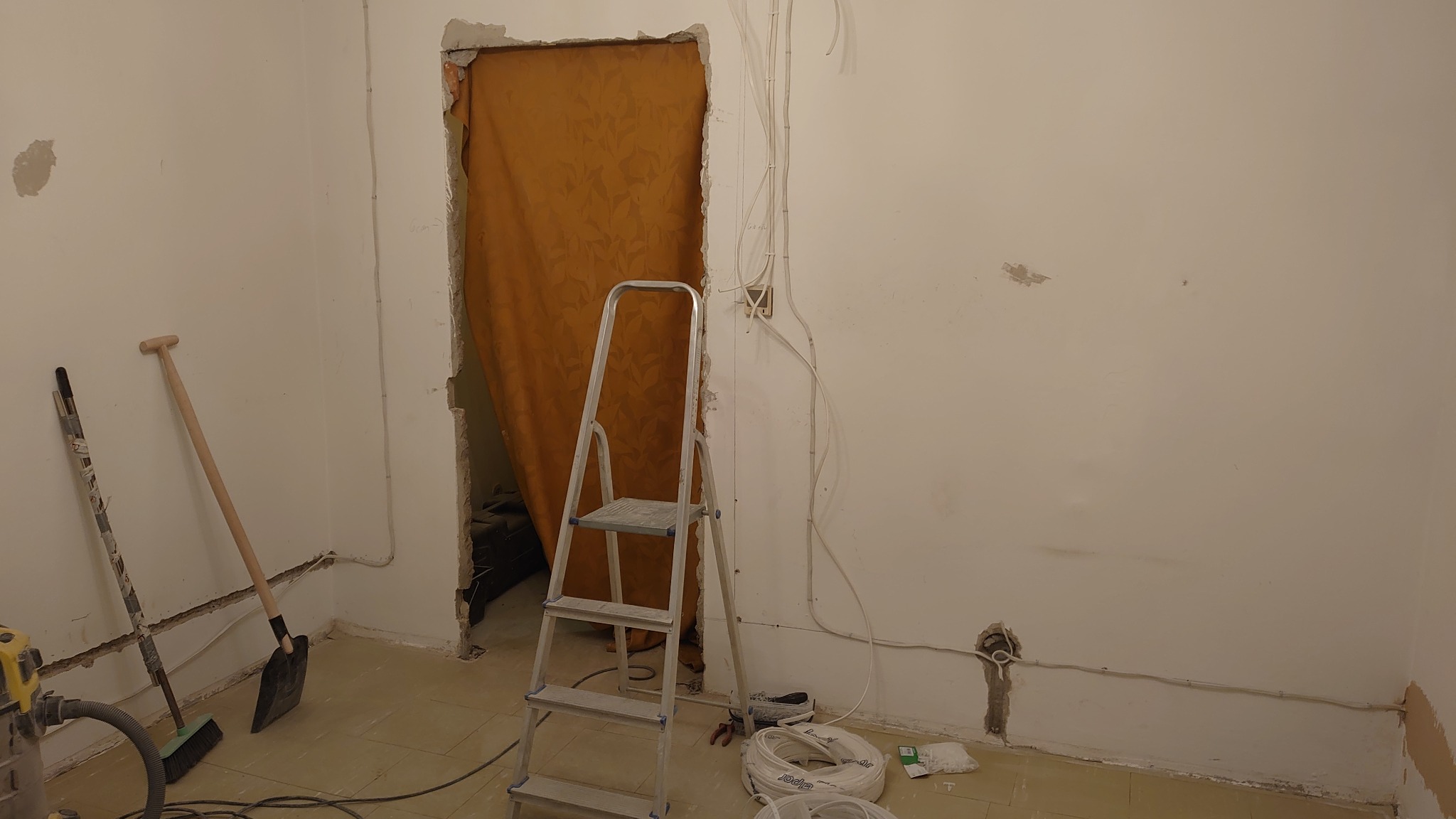 Wnętrze w trakcie remontu: widoczne kable elektryczne na ścianie, drabina, szpachla i łopata. Przez otwór w ścianie widać zasłonę.