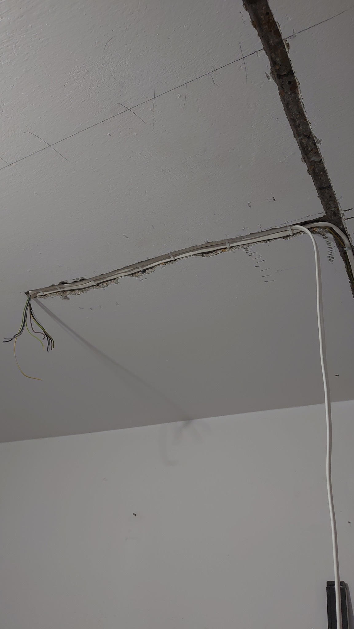 Biały sufit z wyżłobionym kanałem, w którym ułożone są białe przewody. Z kanału wystają kolorowe kable. Widoczne ślady po wierceniu.