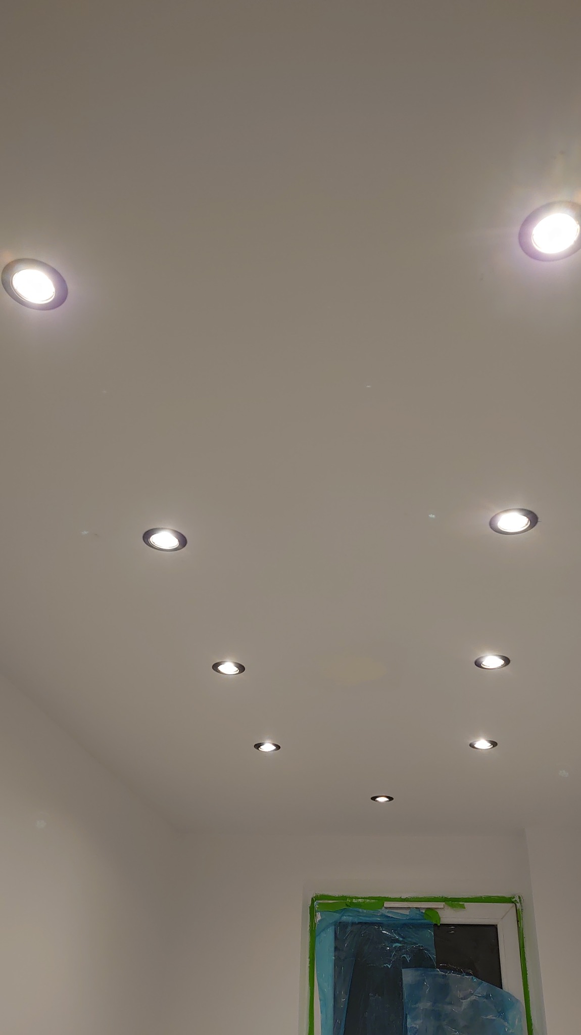 Biały sufit z równomiernie rozmieszczonymi, okrągłymi lampami halogenowymi. Okno zabezpieczone folią malarską w dolnej części kadru.