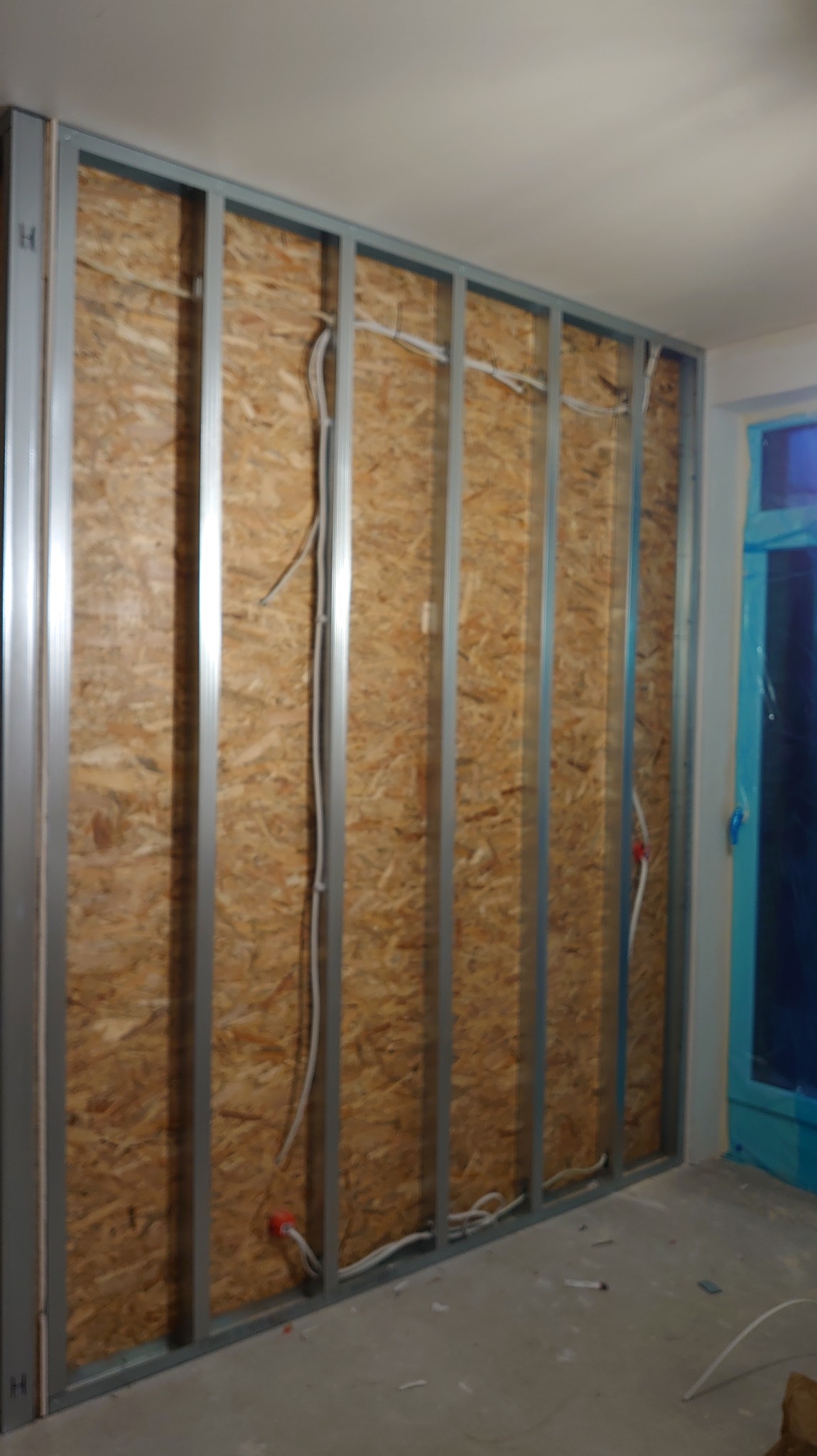 Ściana z płyt OSB i metalowym stelażem, wewnątrz którego poprowadzone są kable elektryczne. Widoczne puszki instalacyjne. Etap prac instalacyjnych.