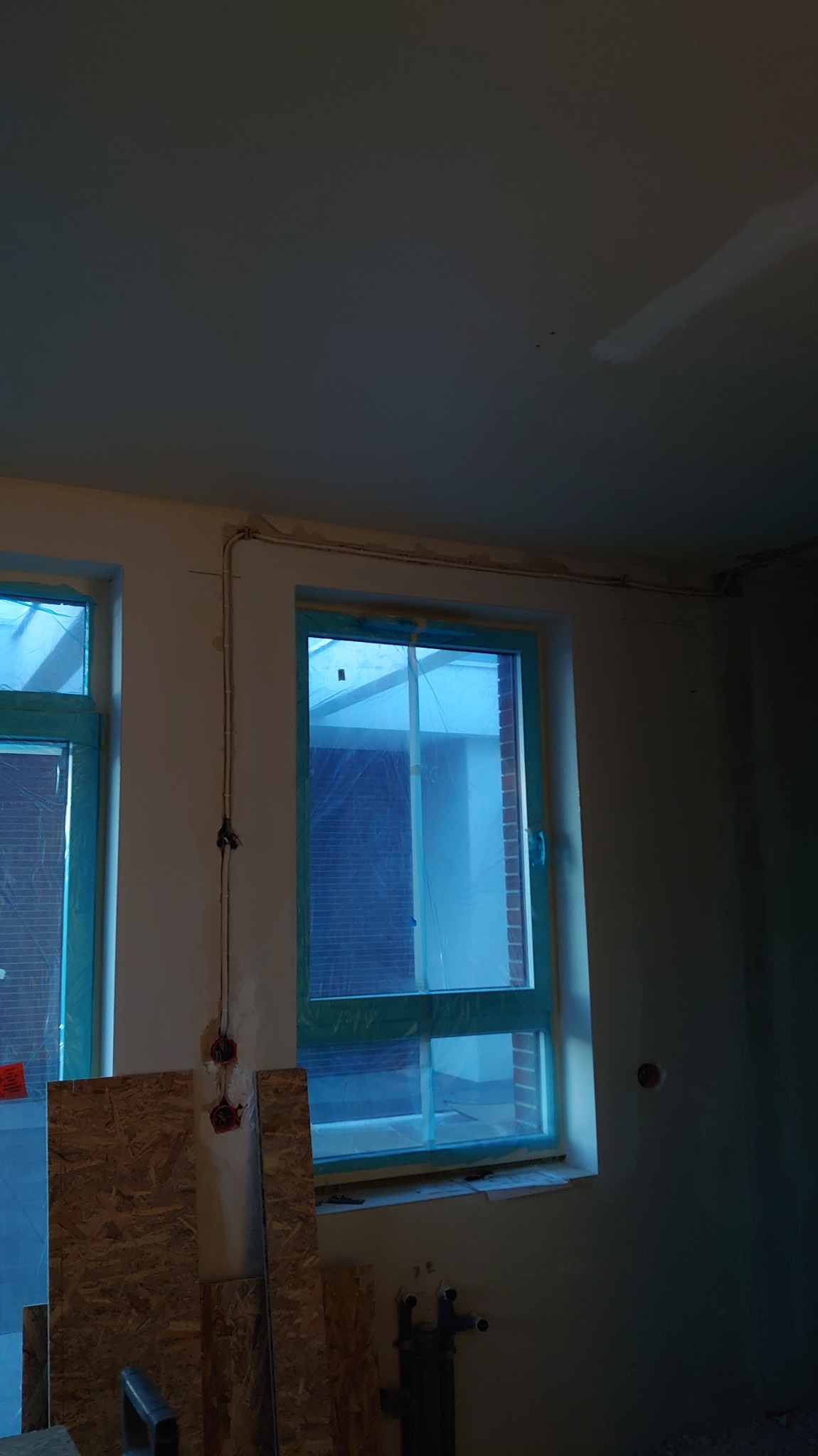 Wnętrze w trakcie remontu: widoczne okna zabezpieczone folią, wystające kable elektryczne i fragmenty płyt OSB. Ściany przygotowane do dalszych prac wykończeniowych.