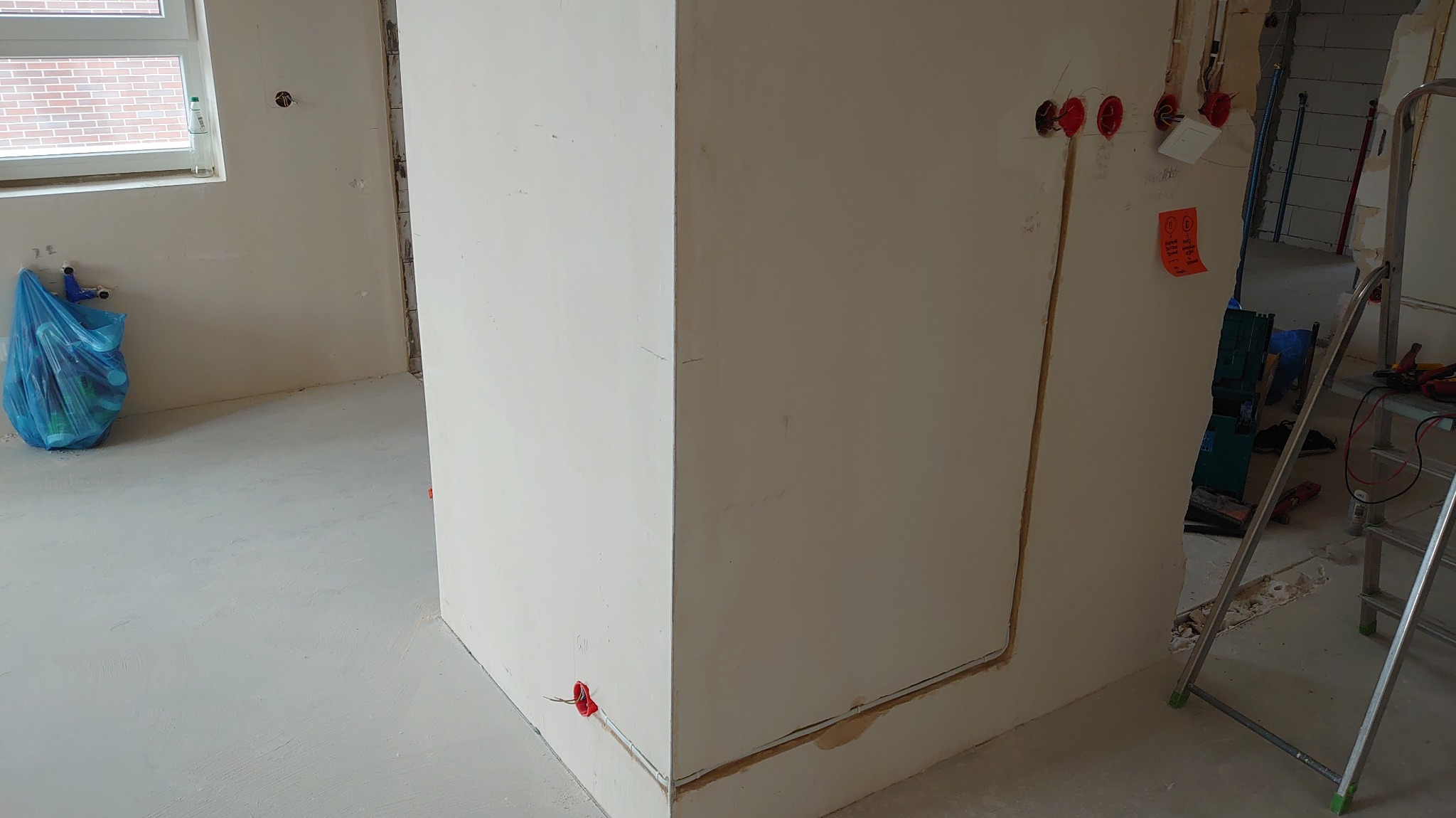 Ściana z widocznymi puszkami instalacyjnymi i przewodami w trakcie prac wykończeniowych. W tle drabina i narzędzia.