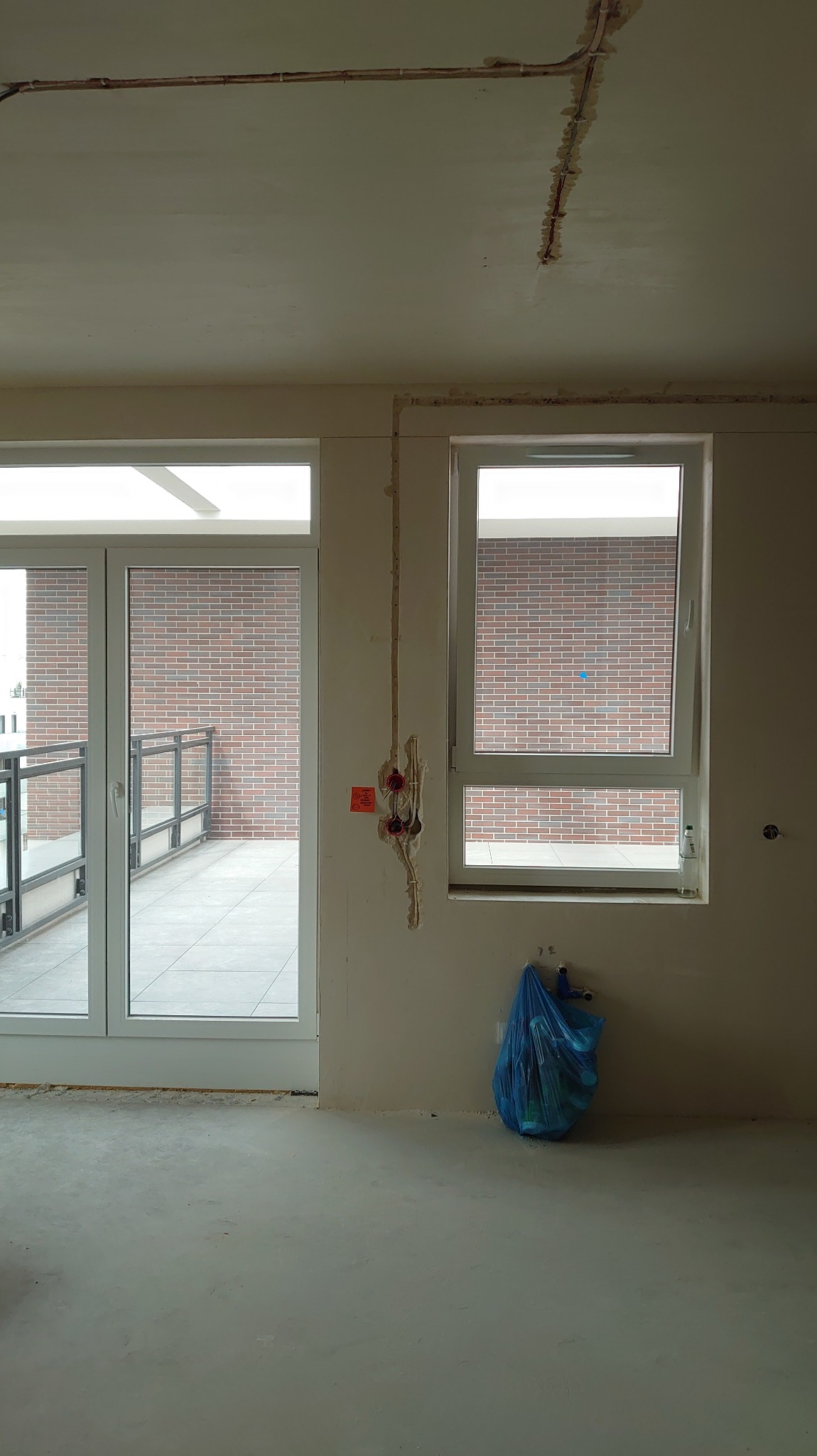 Wnętrze w stanie deweloperskim z widocznymi puszkami elektrycznymi i przewodami na ścianie, okno z widokiem na ceglany budynek, worek z odpadami budowlanymi.