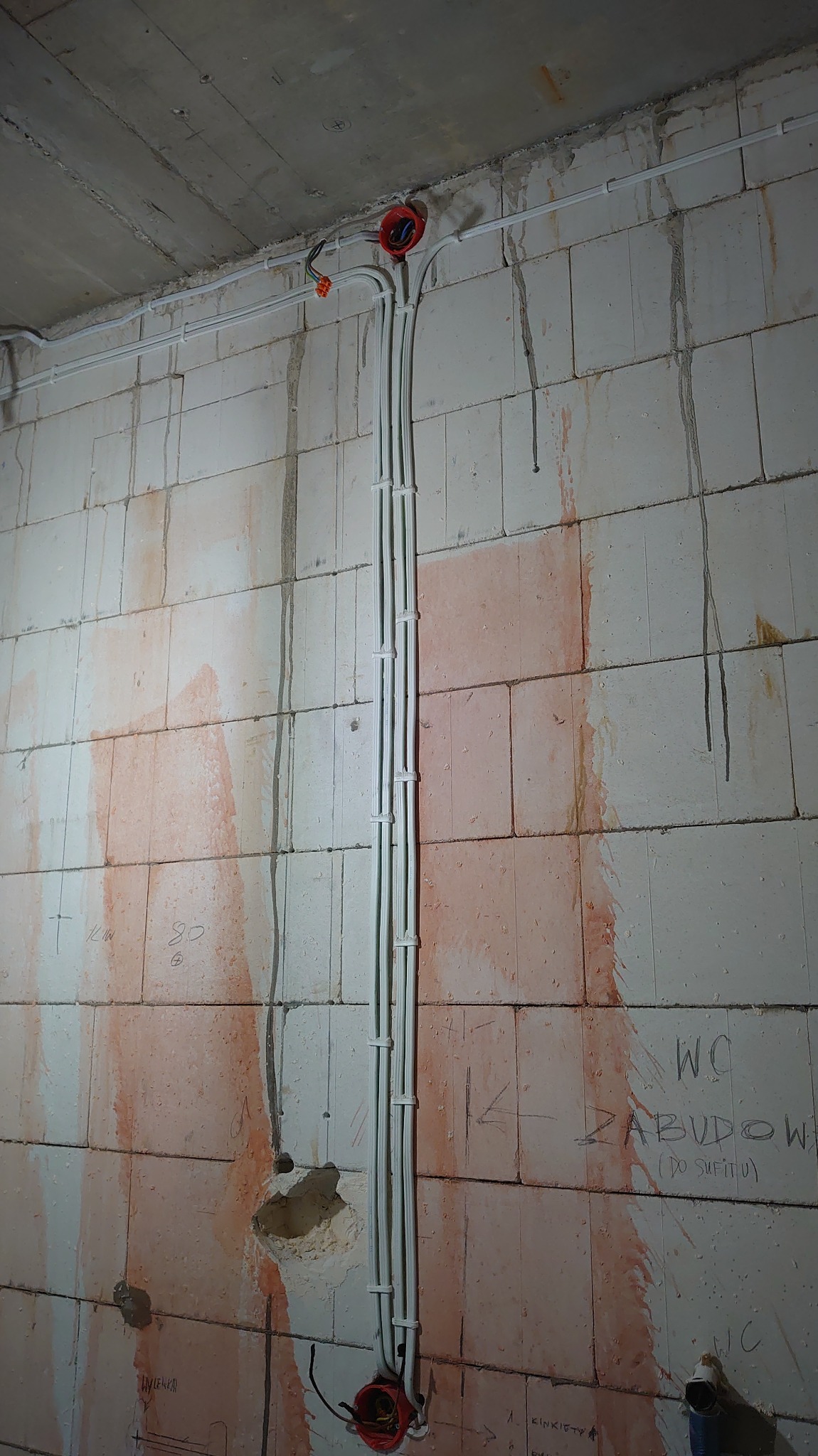 Surowa ściana z betonu komórkowego z pionowo poprowadzonymi, białymi rurkami instalacji elektrycznej, zakończonymi czerwonymi puszkami. Widoczne ślady po instalacji.