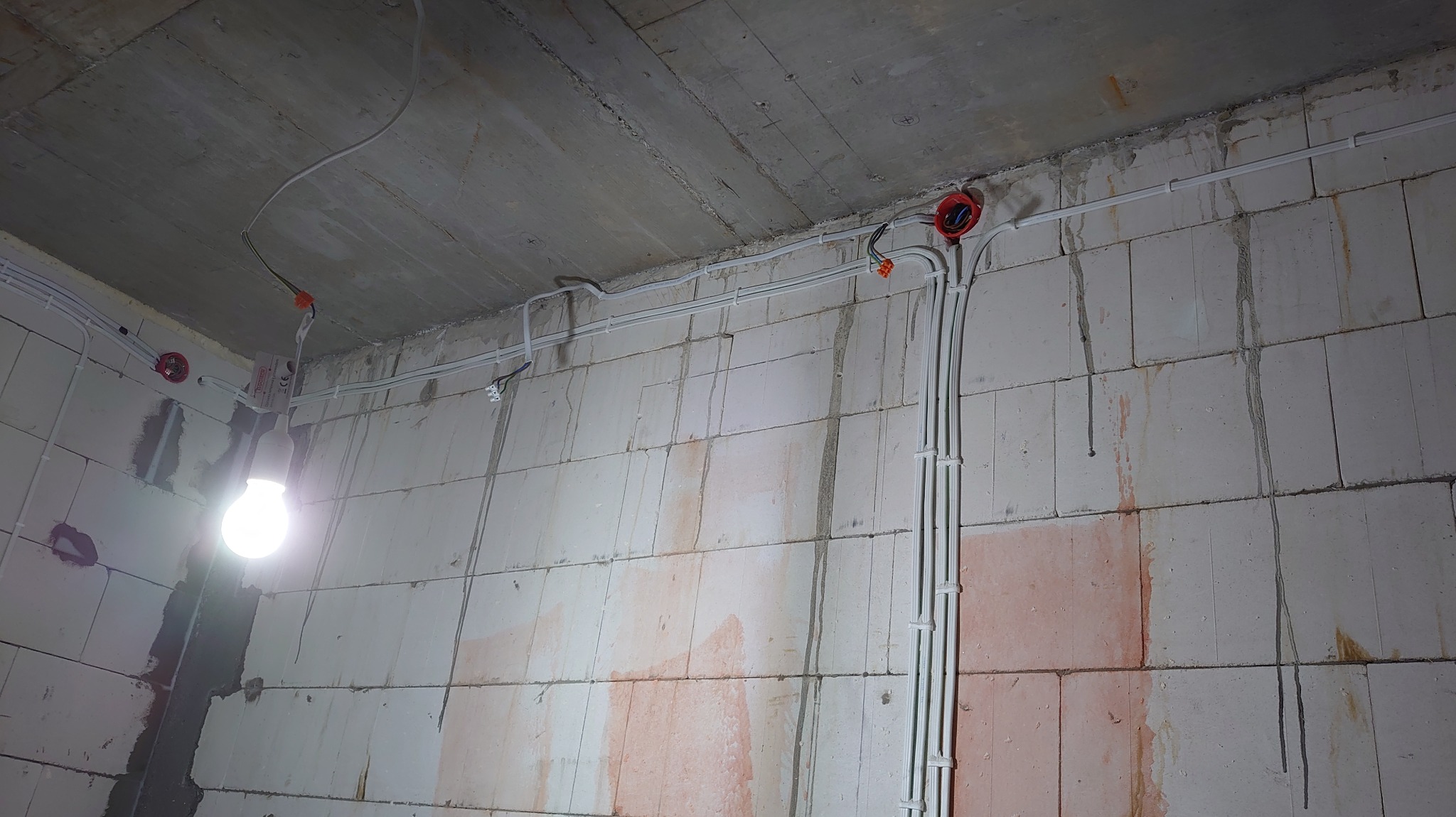 Surowy mur z widocznymi przewodami w białych listwach, puszkami instalacyjnymi i żarówką. Instalacja w trakcie realizacji, widoczne ślady po pracach budowlanych.