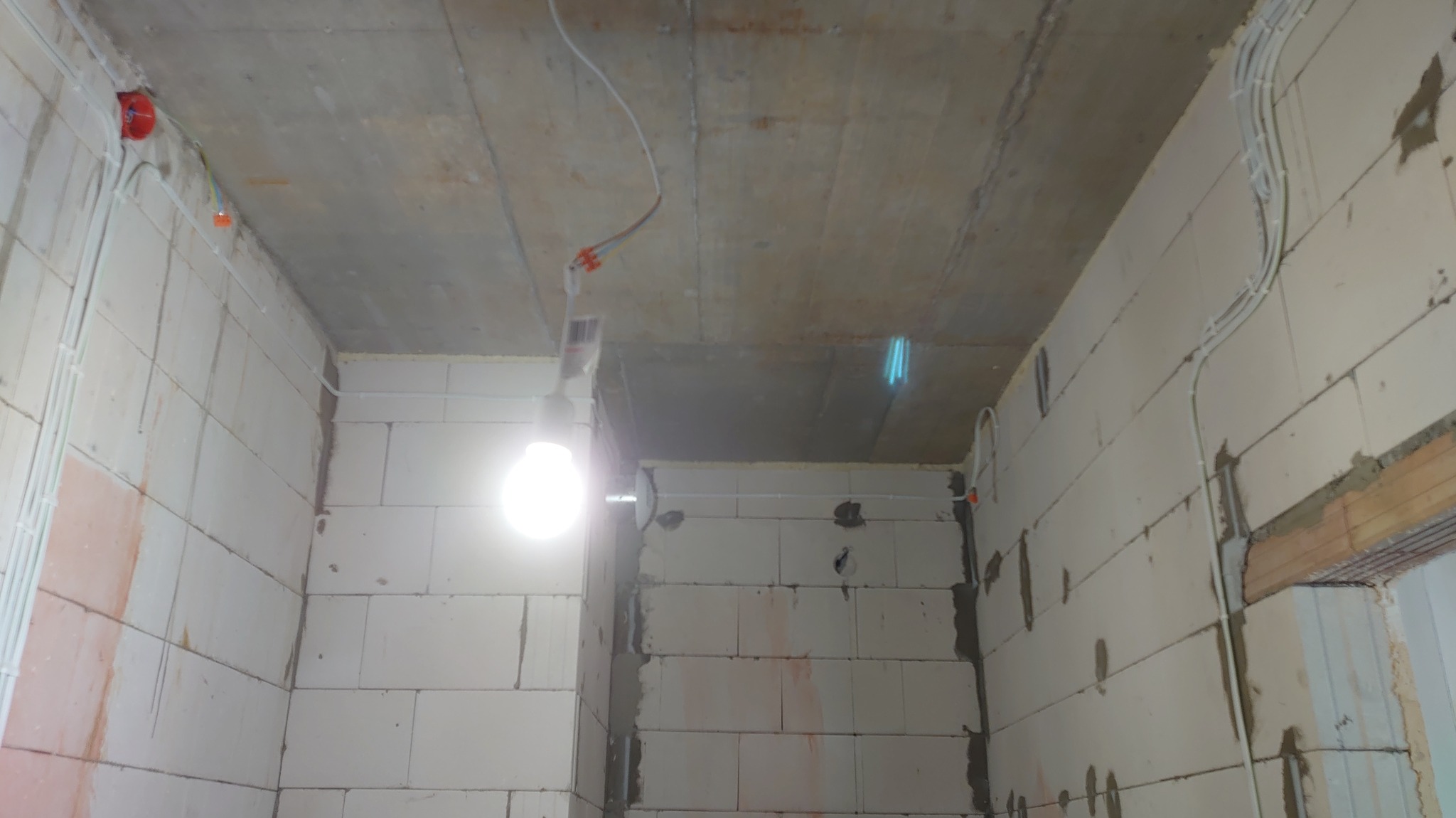 Wnętrze w trakcie prac wykończeniowych z widoczną instalacją elektryczną w peszlach na ścianach z betonu komórkowego i wiszącą żarówką.