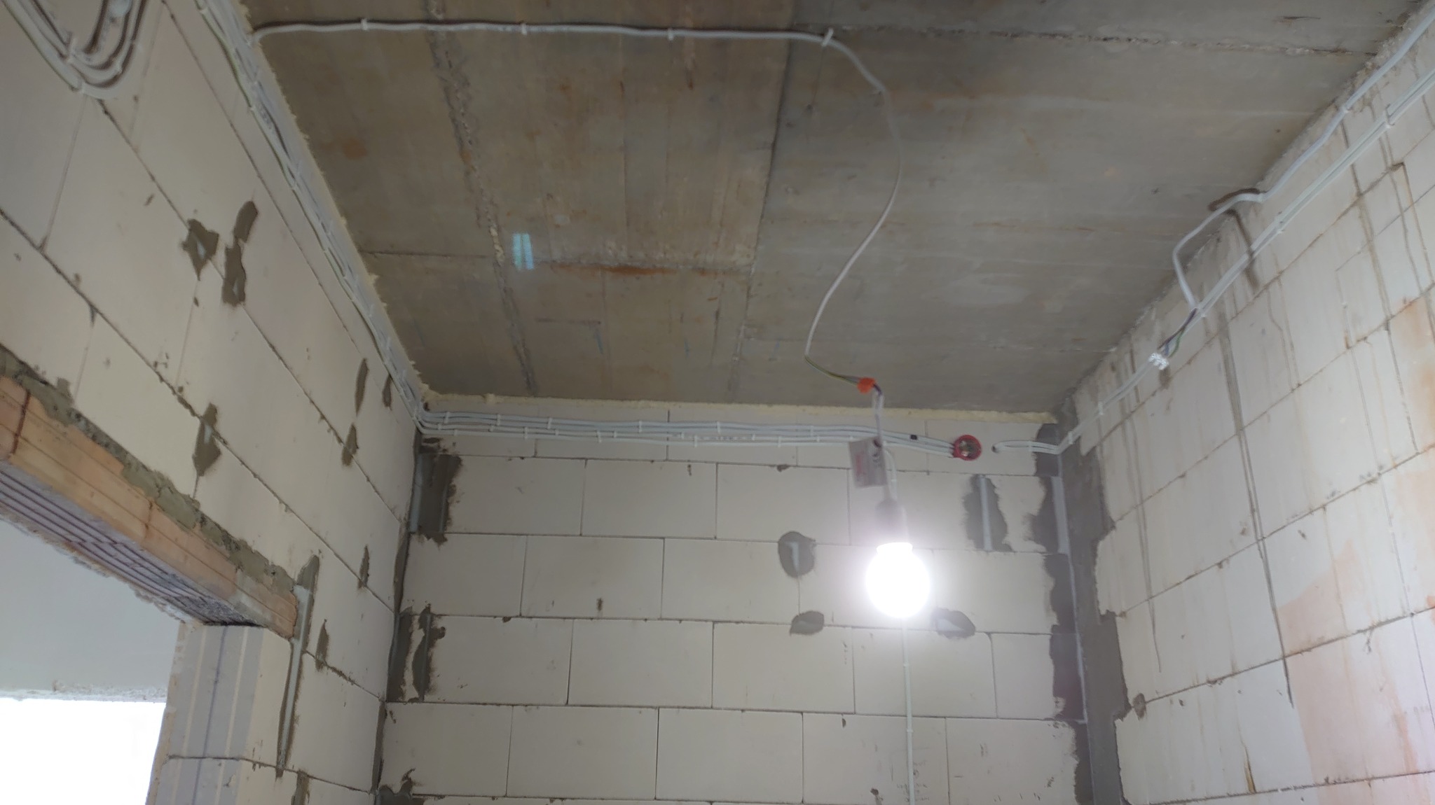 Wnętrze w trakcie budowy z widocznymi przewodami elektrycznymi ułożonymi wzdłuż ścian i sufitu, z żarówką tymczasową. Surowy stan wykończenia.