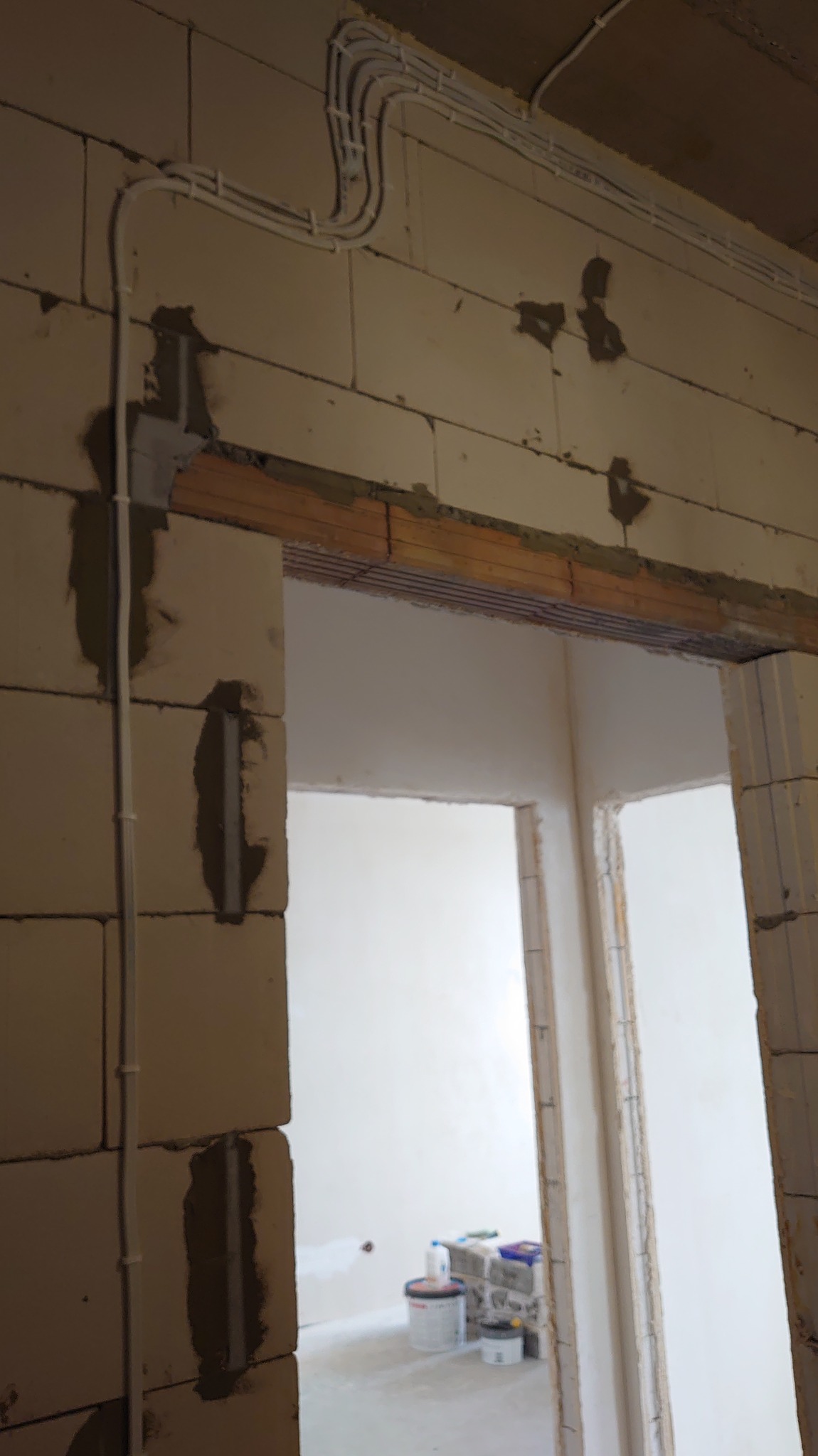 Białe rury instalacji elektrycznej prowadzone po ścianie z betonu komórkowego nad otworem drzwiowym. Widoczne ślady po montażu i zaprawa. W tle widok na jasne pomieszczenie.