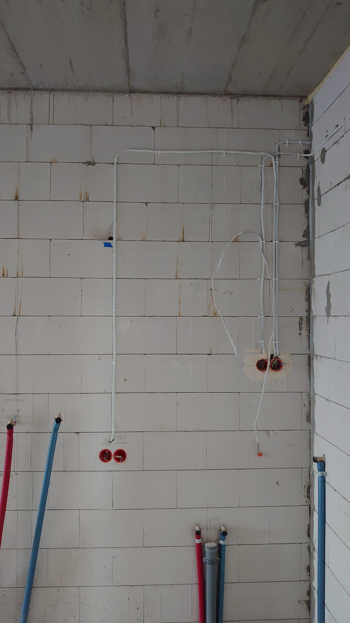 Surowa ściana z betonu komórkowego z widocznymi przewodami elektrycznymi i puszkami instalacyjnymi. Instalacja w trakcie montażu, widoczne przewody i rury.