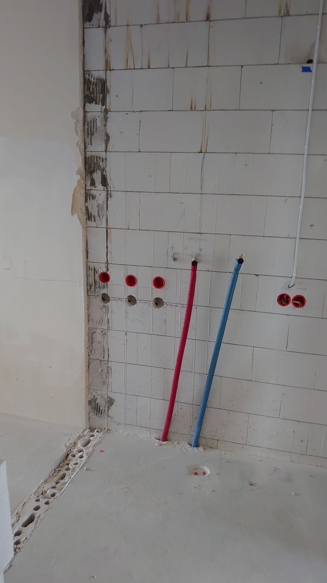 Instalacja elektryczna i hydrauliczna w trakcie remontu: widoczne rury (czerwona i niebieska) oraz puszki elektryczne w ścianie z białej cegły.