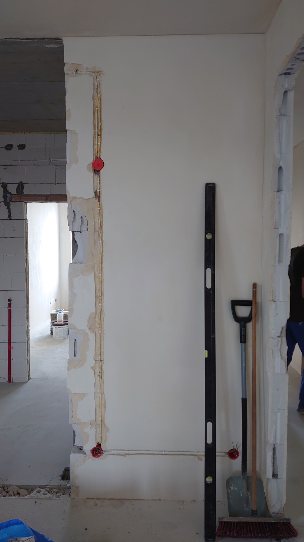 Wnętrze w trakcie remontu: widoczne kable elektryczne w ścianie, poziomica, szpachelka i zmiotka. Instalacja w trakcie wykonywania, surowy stan wykończenia.